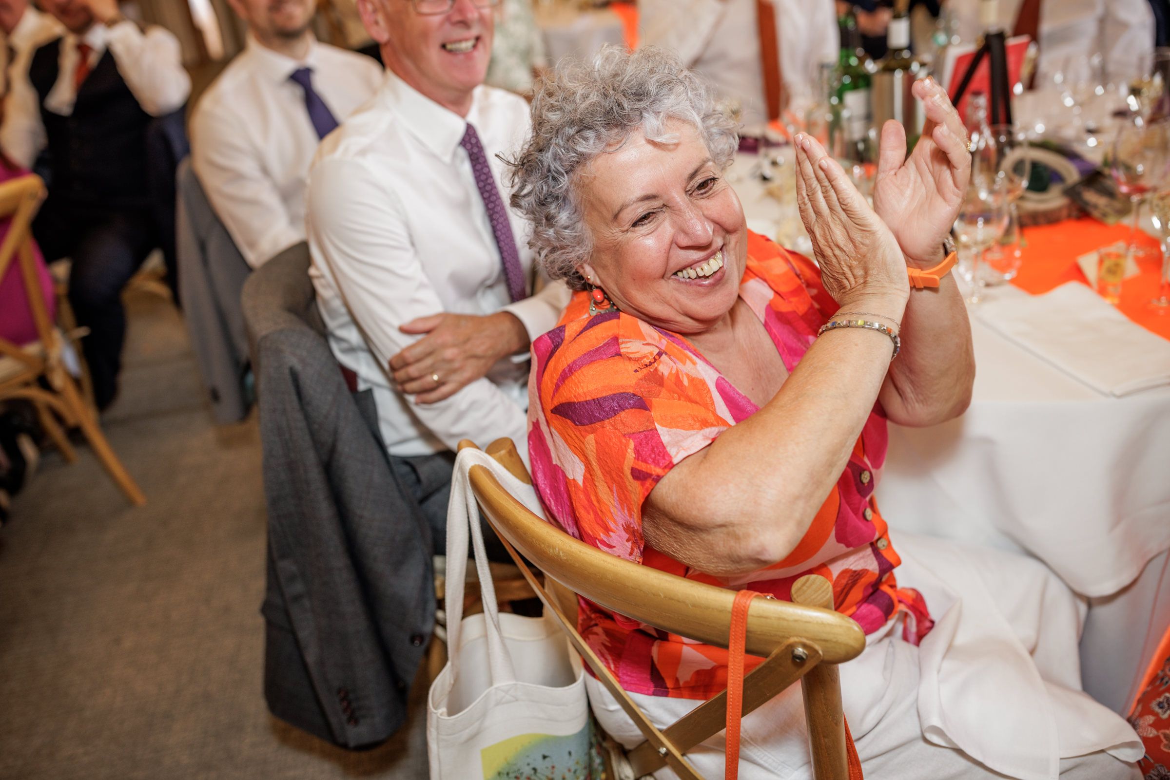 Elderly woman in colorful pink dress laughing and clapping at a wedding reception celebration.