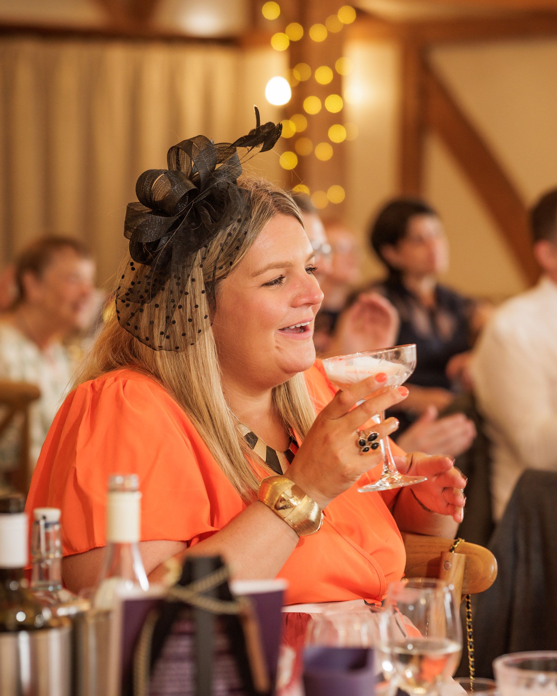 Woman in orange dress and black fascinator laughs while holding a cocktail glass at an elegant indoor event.