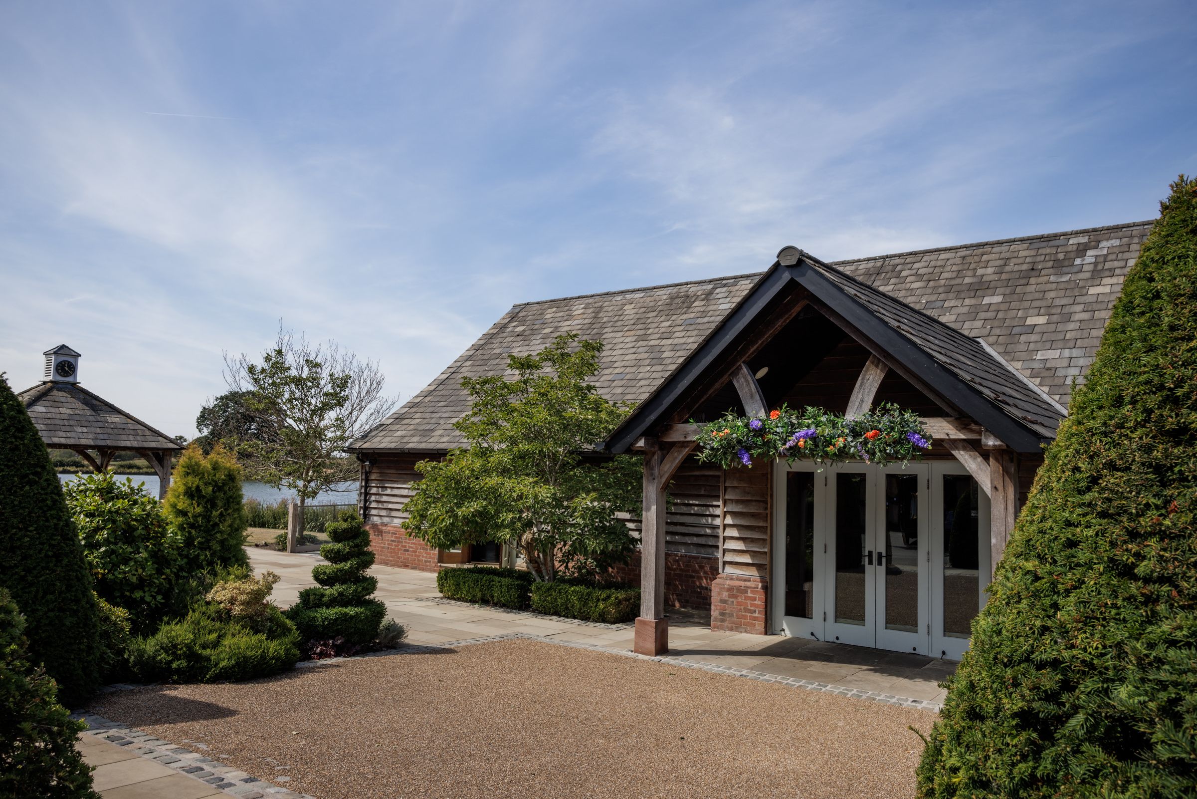 Charming countryside barn venue with timber-framed porch, hanging flowers, slate roof, and manicured hedges under a blue sky.