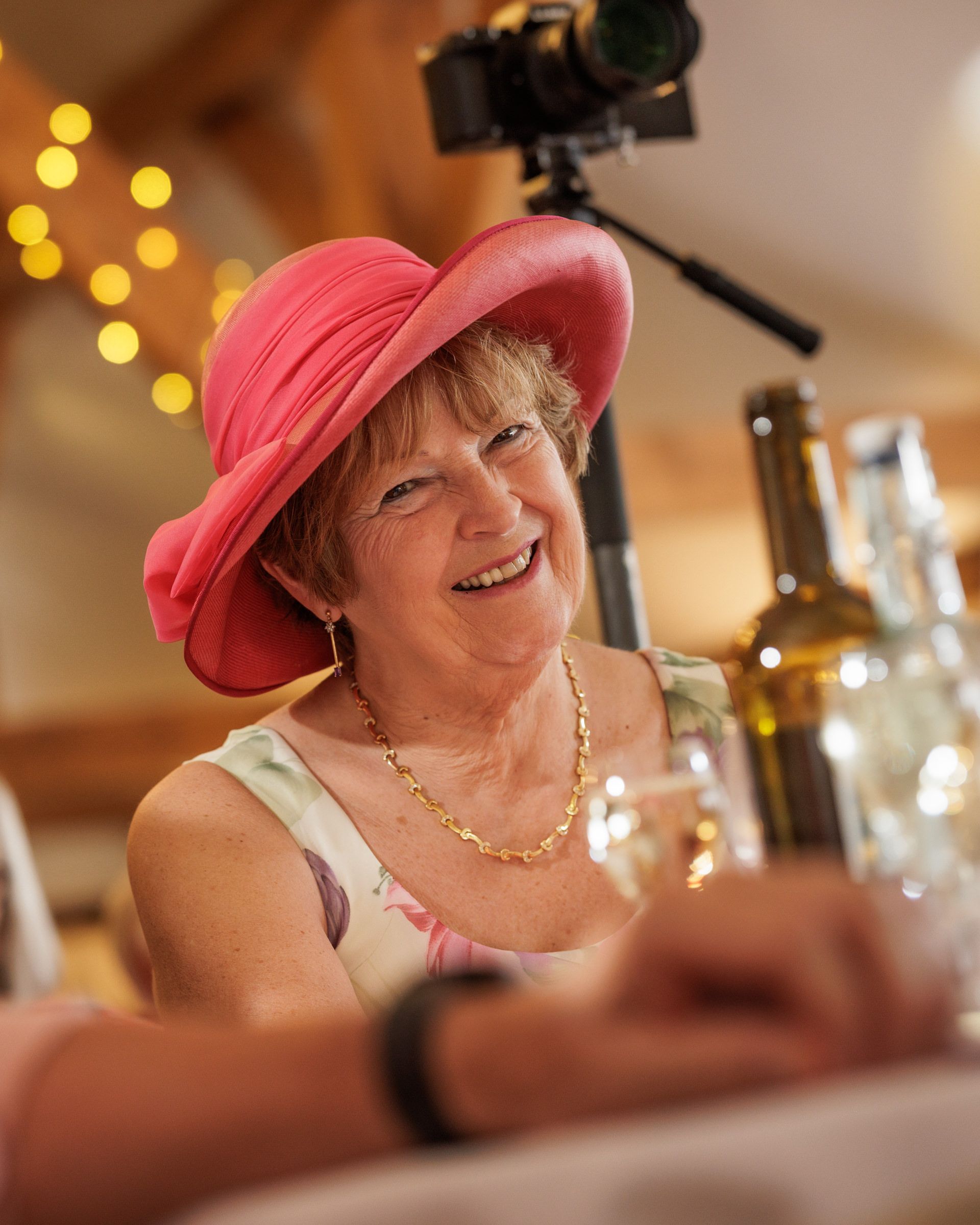 Smiling older woman wearing a pink wide-brim hat and gold necklace at an indoor event with bokeh lights.