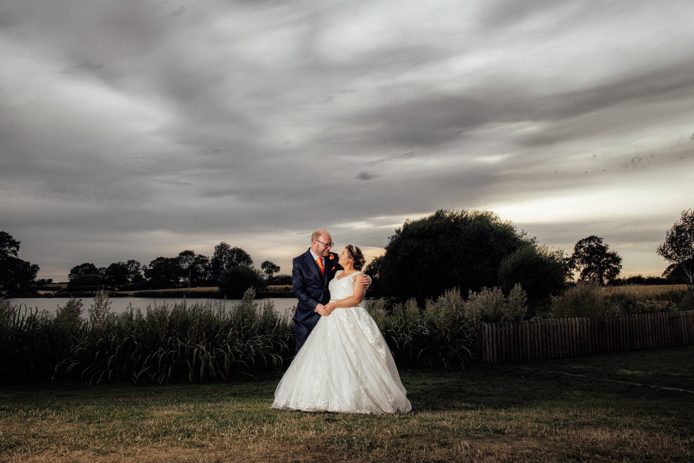Bride and groom embrace near a lakeside at dusk under dramatic cloudy skies in an outdoor wedding portrait.