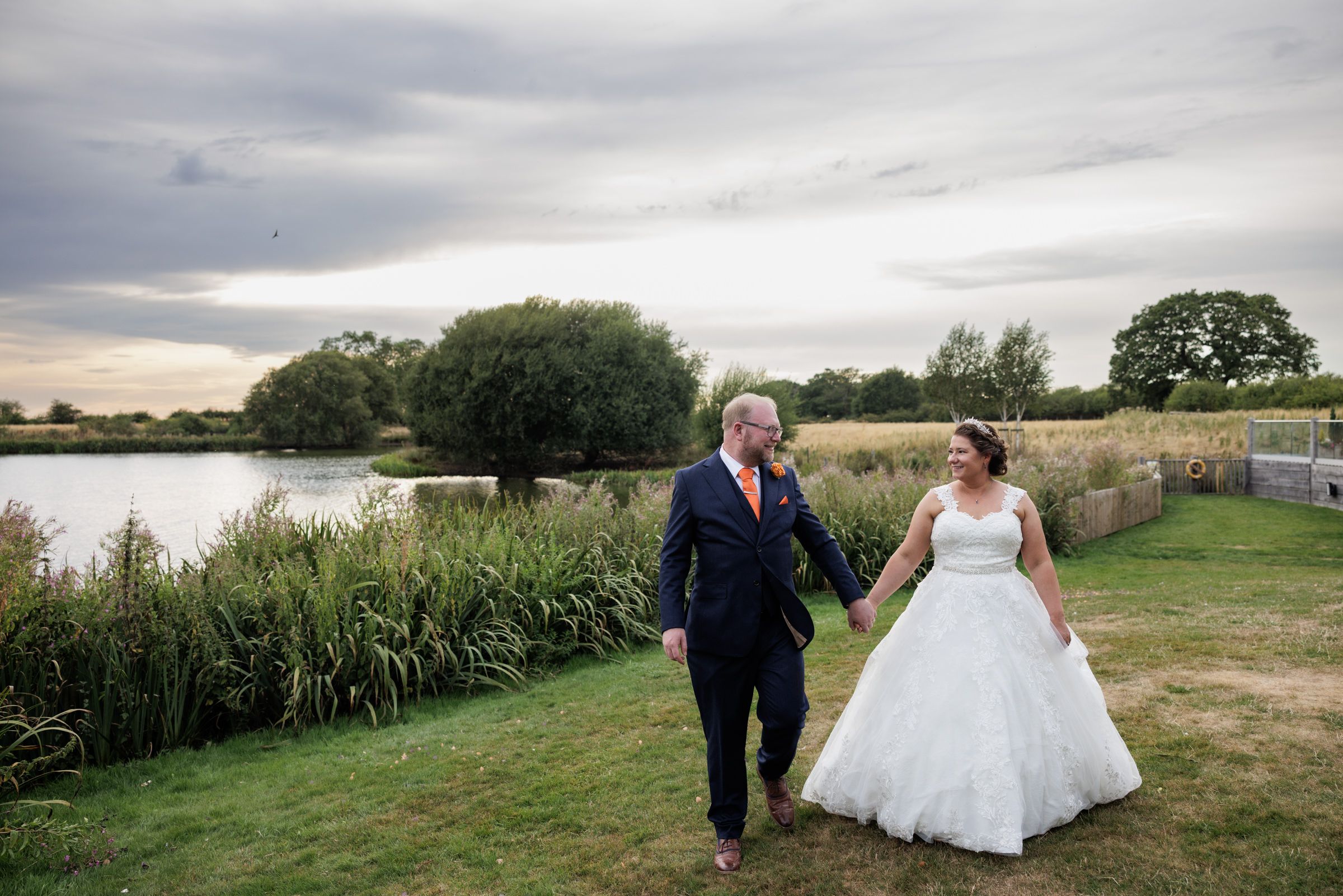 Bride and groom walk hand-in-hand by a lakeside, smiling at each other in a lush green outdoor wedding setting.