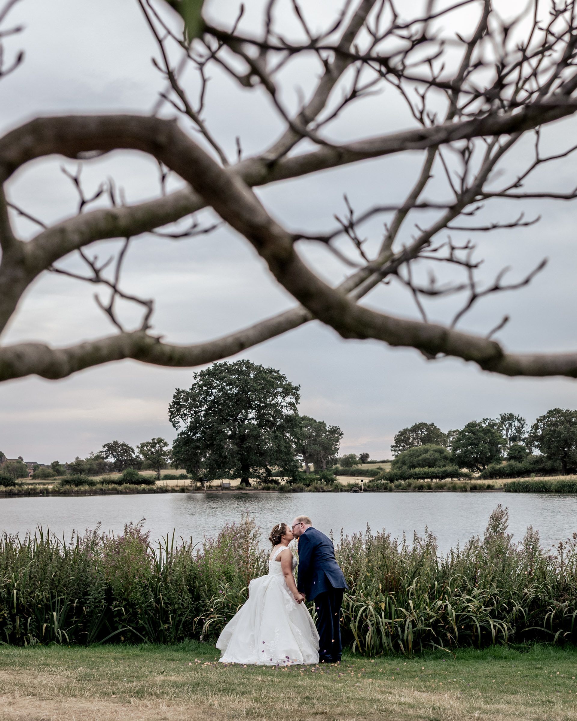 A bride and groom share a kiss by a lakeside, framed by bare tree branches against a cloudy sky.