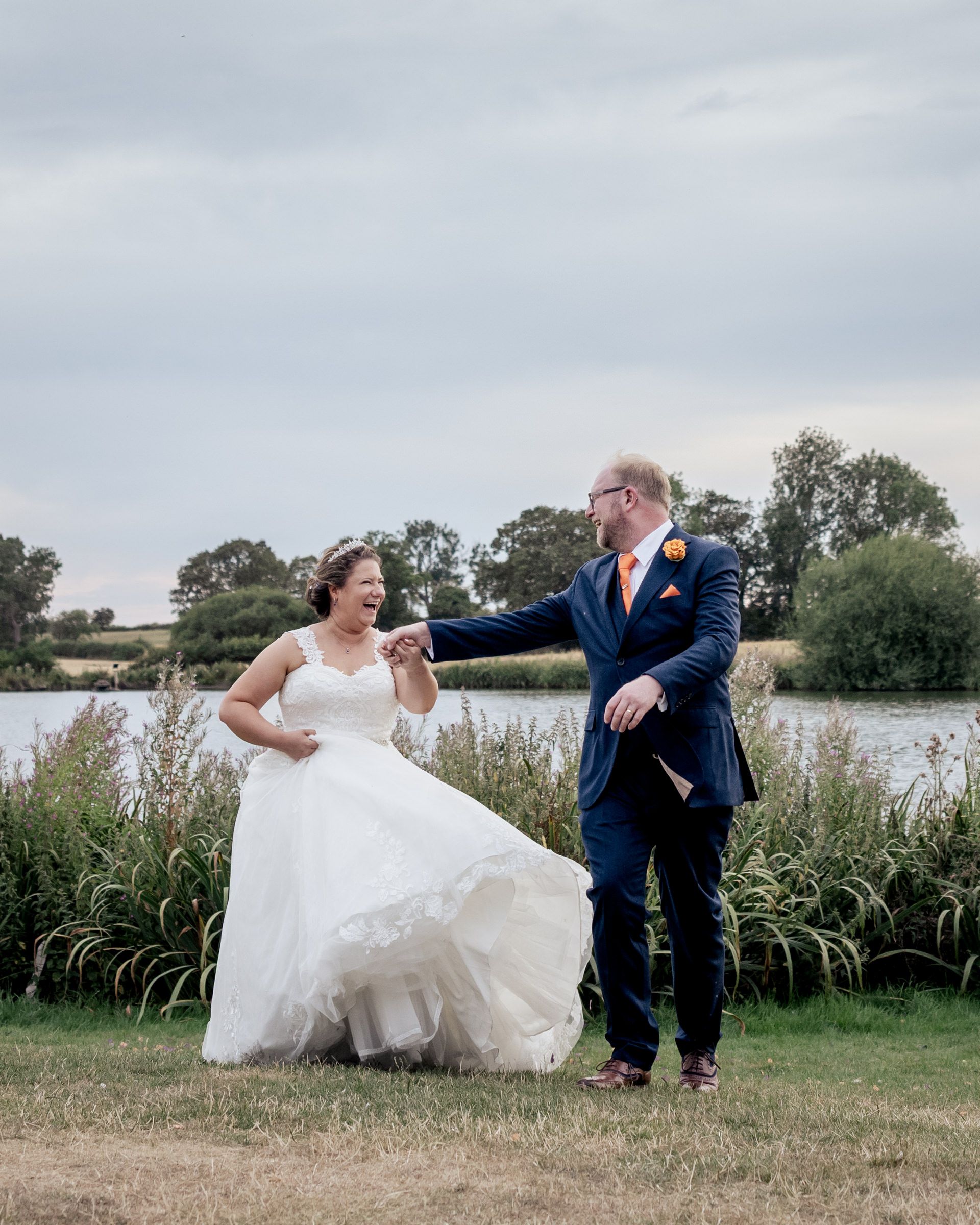 A bride in a white gown and groom in a navy suit hold hands by a lakeside, sharing a joyful wedding moment.