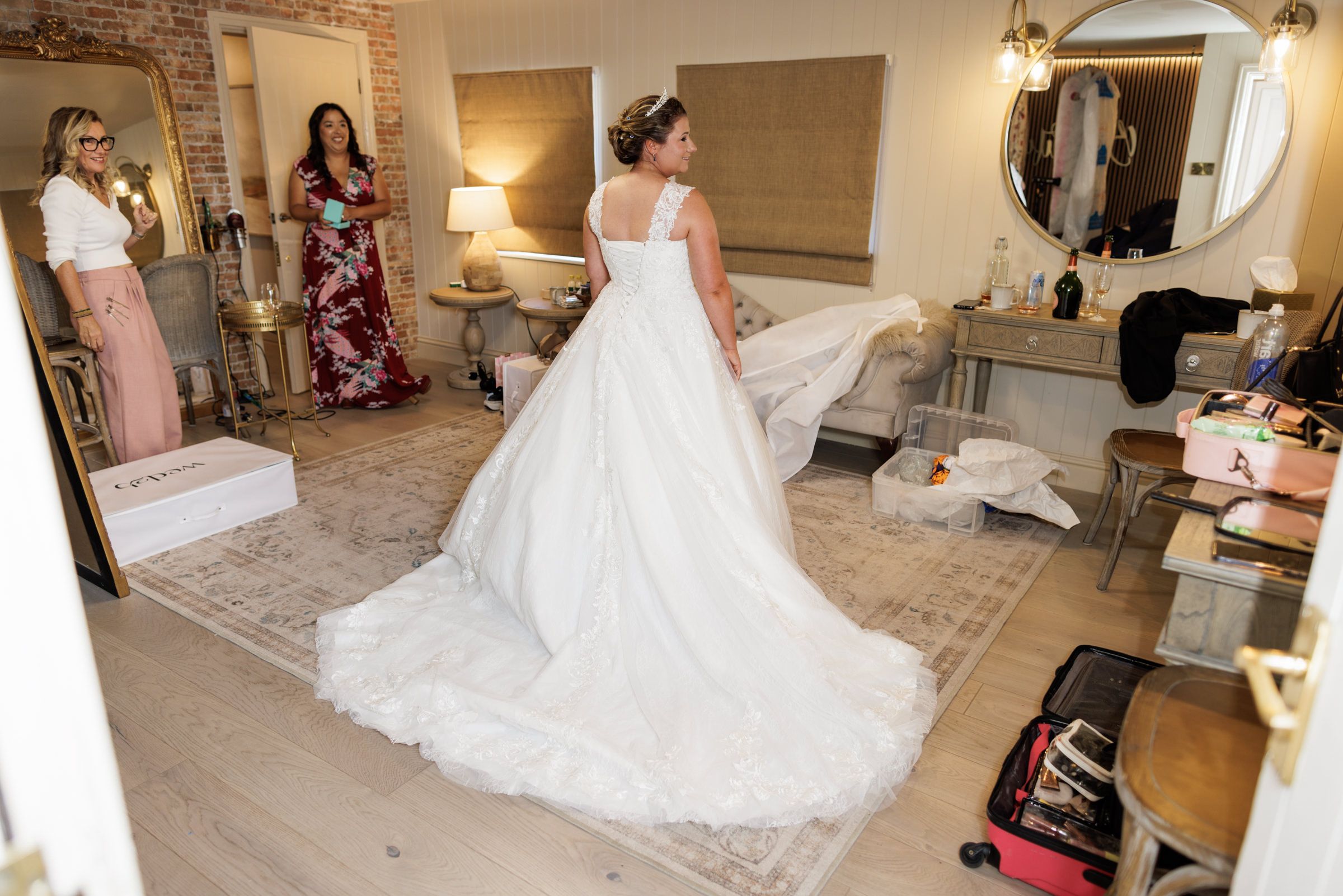 Bride in elegant white ball gown with long train stands in dressing room as bridesmaids look on admiringly.