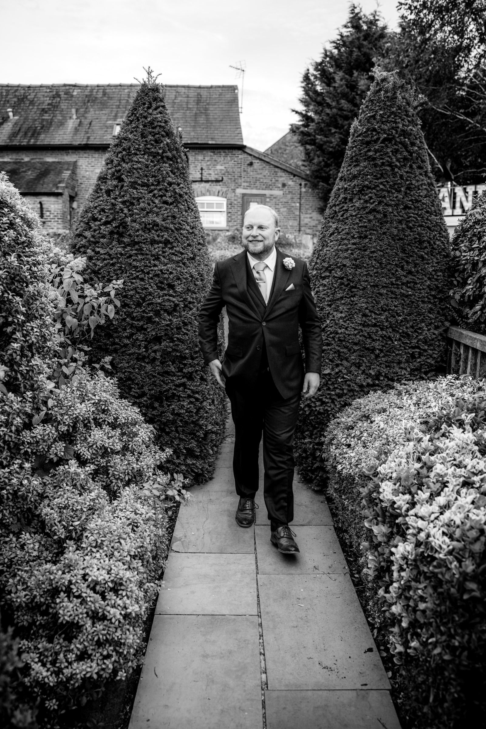A groom in a dark suit walks along a garden path flanked by tall topiary trees at a wedding venue.