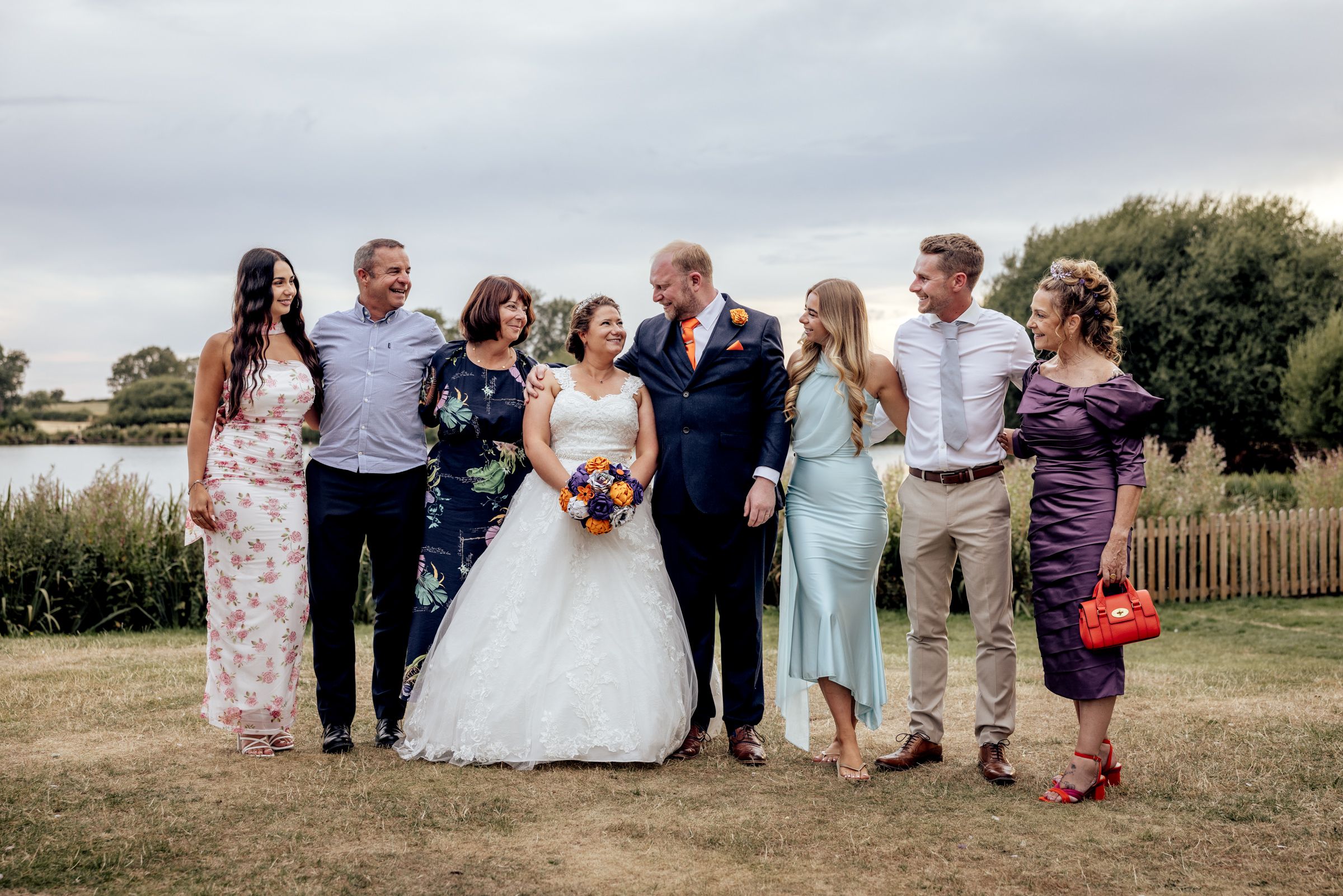 Wedding party poses outdoors by a lake, bride in white gown holding orange bouquet with family in formal attire.