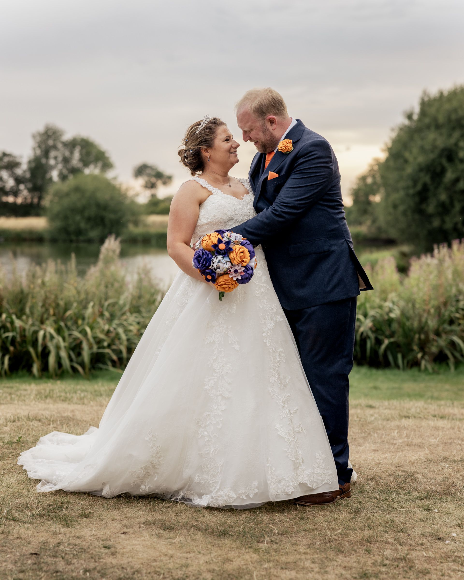Bride and groom share romantic moment outdoors, she in lace ballgown holding purple orange bouquet, he in navy suit.
