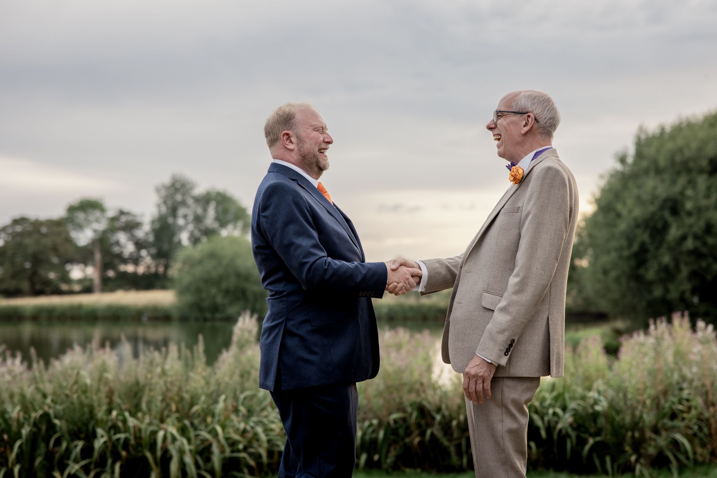 Two men in suits shaking hands outdoors near a pond with lush greenery and a cloudy sky backdrop.
