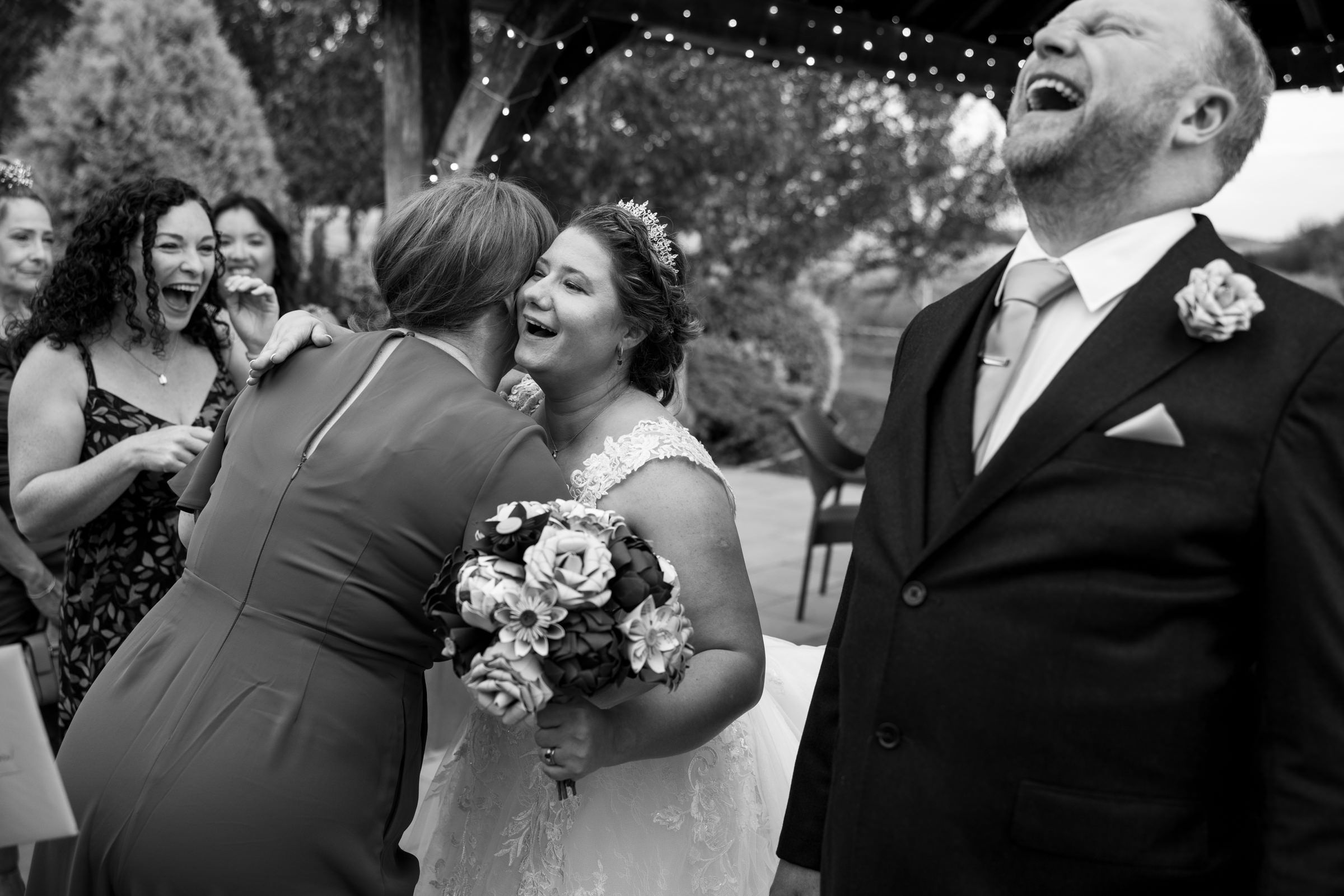Black and white wedding photo of laughing bride, groom, and guests sharing joyful moment after ceremony.