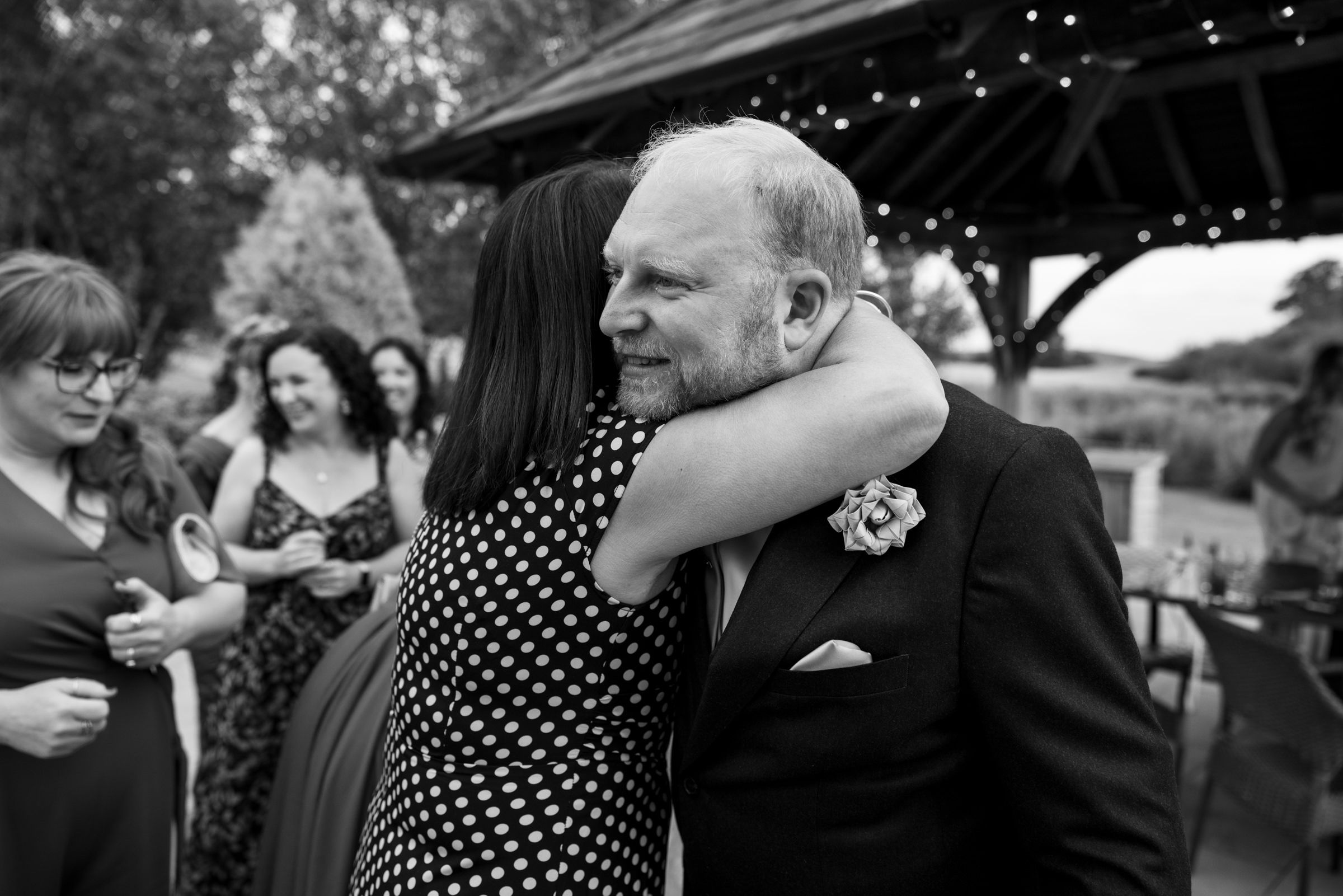 Black and white photo of a woman in polka dot dress hugging an older man in a suit at an outdoor wedding reception.