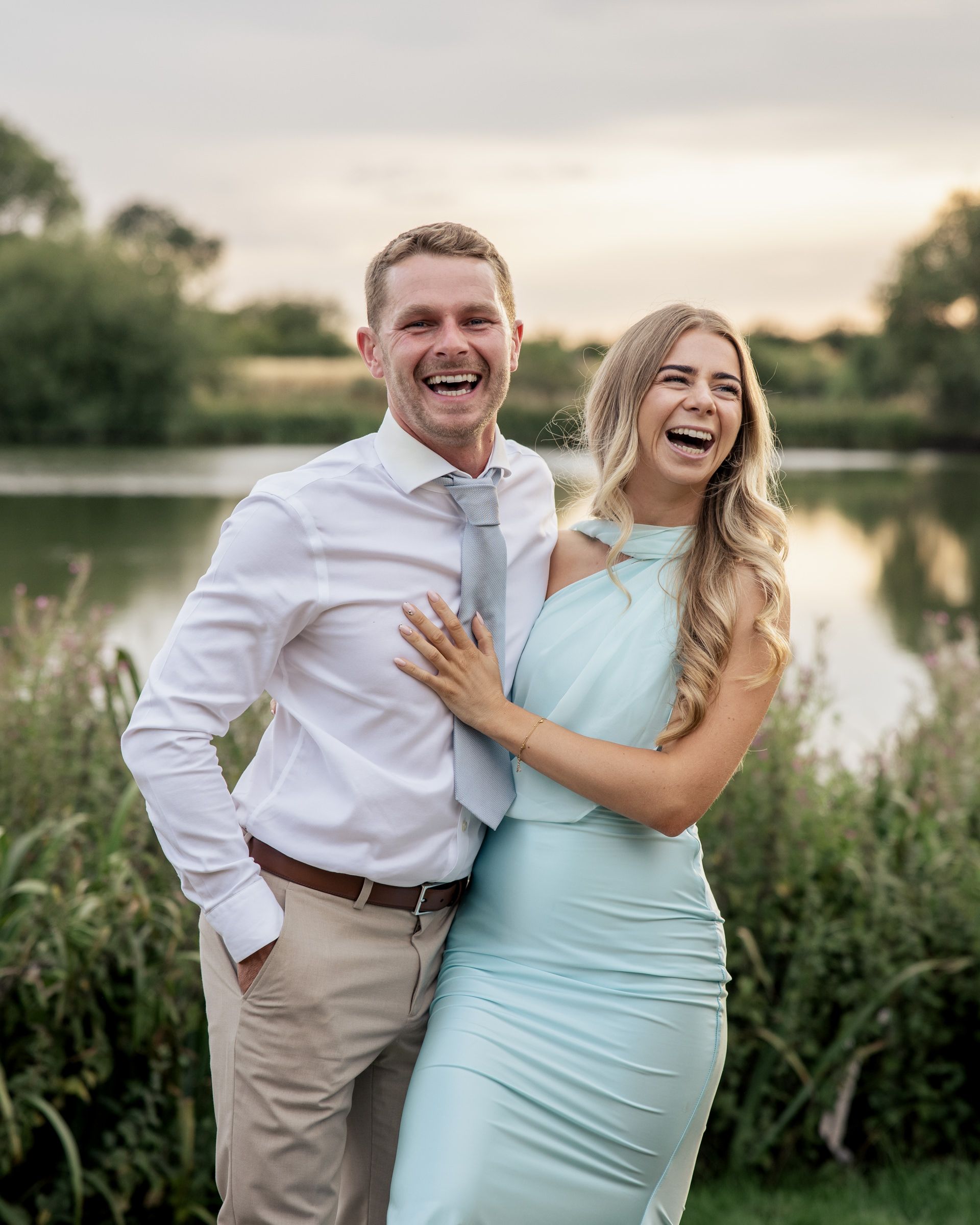 A laughing couple poses outdoors near a lake, man in white shirt and woman in mint dress, during golden hour.
