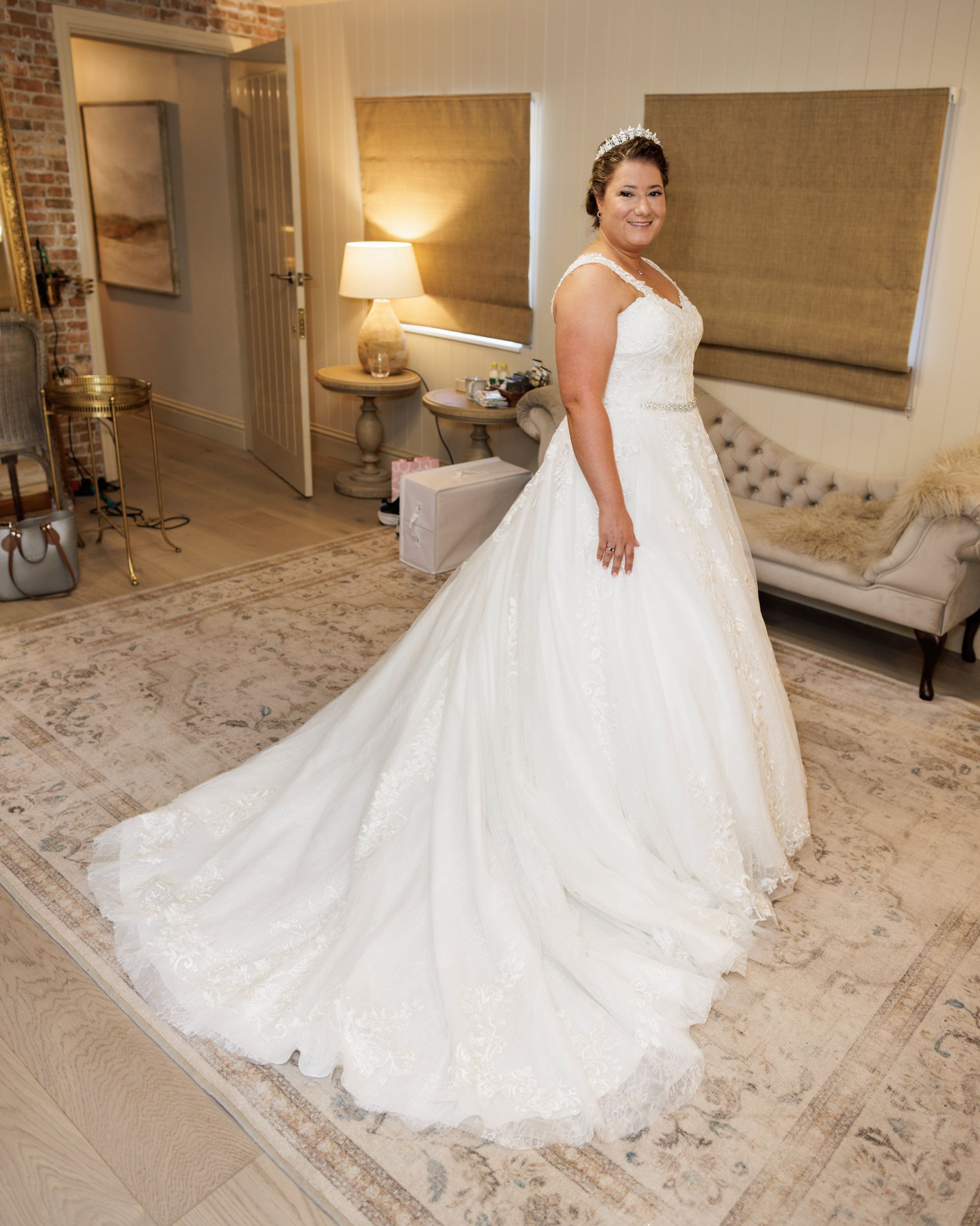 Bride wearing a white A-line wedding gown with lace train and crystal tiara, posing in an elegant room.