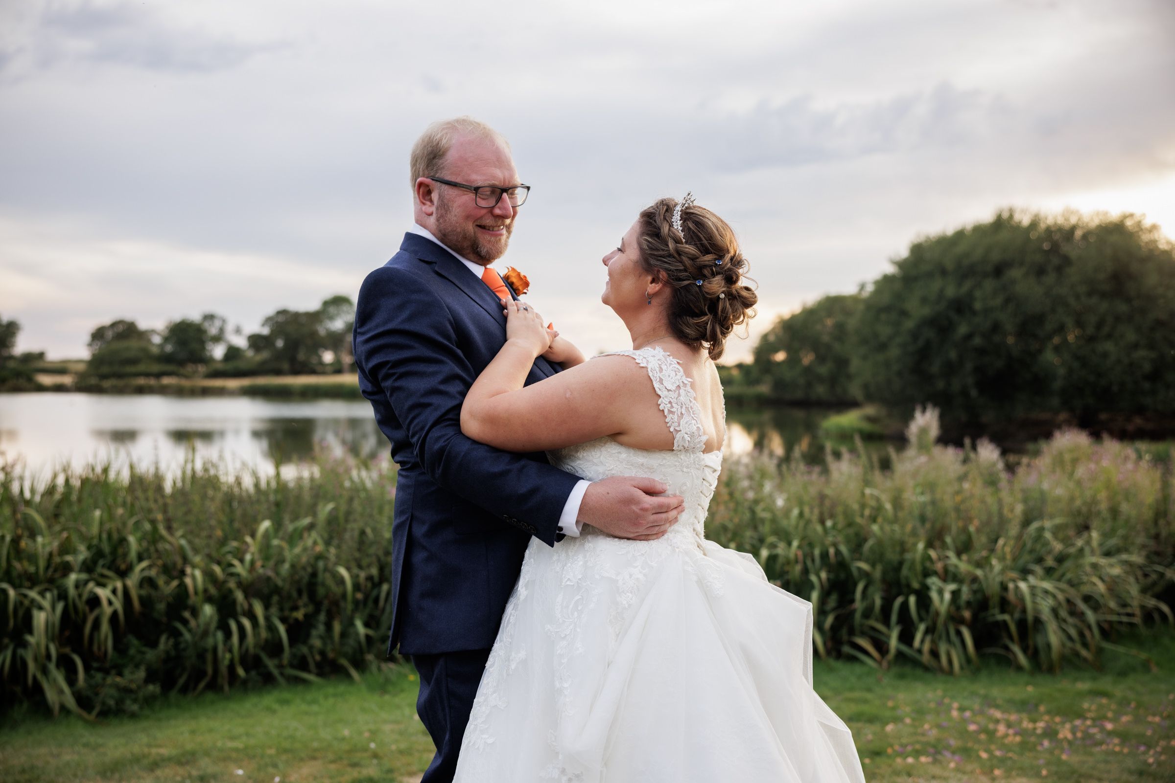 Bride and groom share a romantic dance outdoors near a lake, surrounded by lush greenery at sunset.