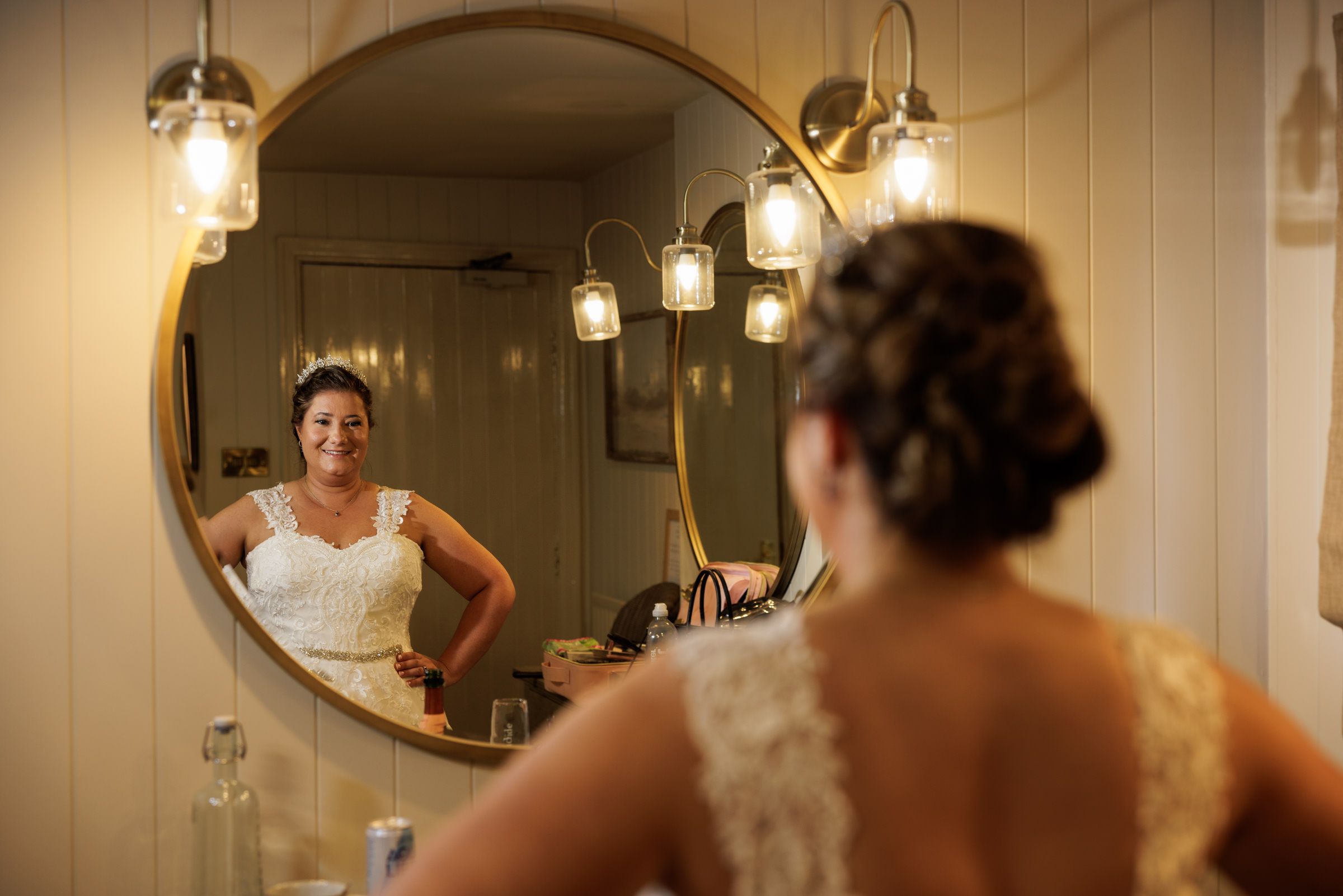 Bride in white lace wedding dress admires her reflection in a round gold-framed mirror with warm pendant lights.