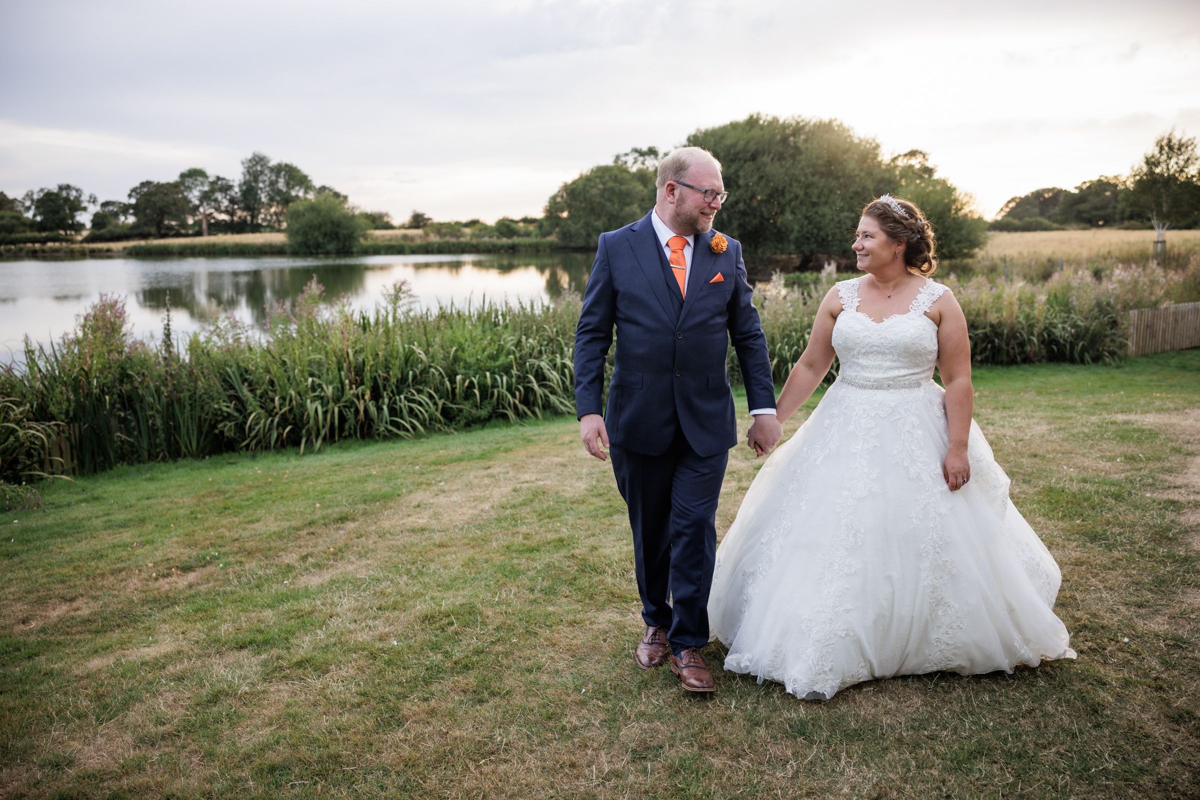 Bride and groom walk hand-in-hand by a serene lake, wearing a white ballgown and navy suit with coral tie.