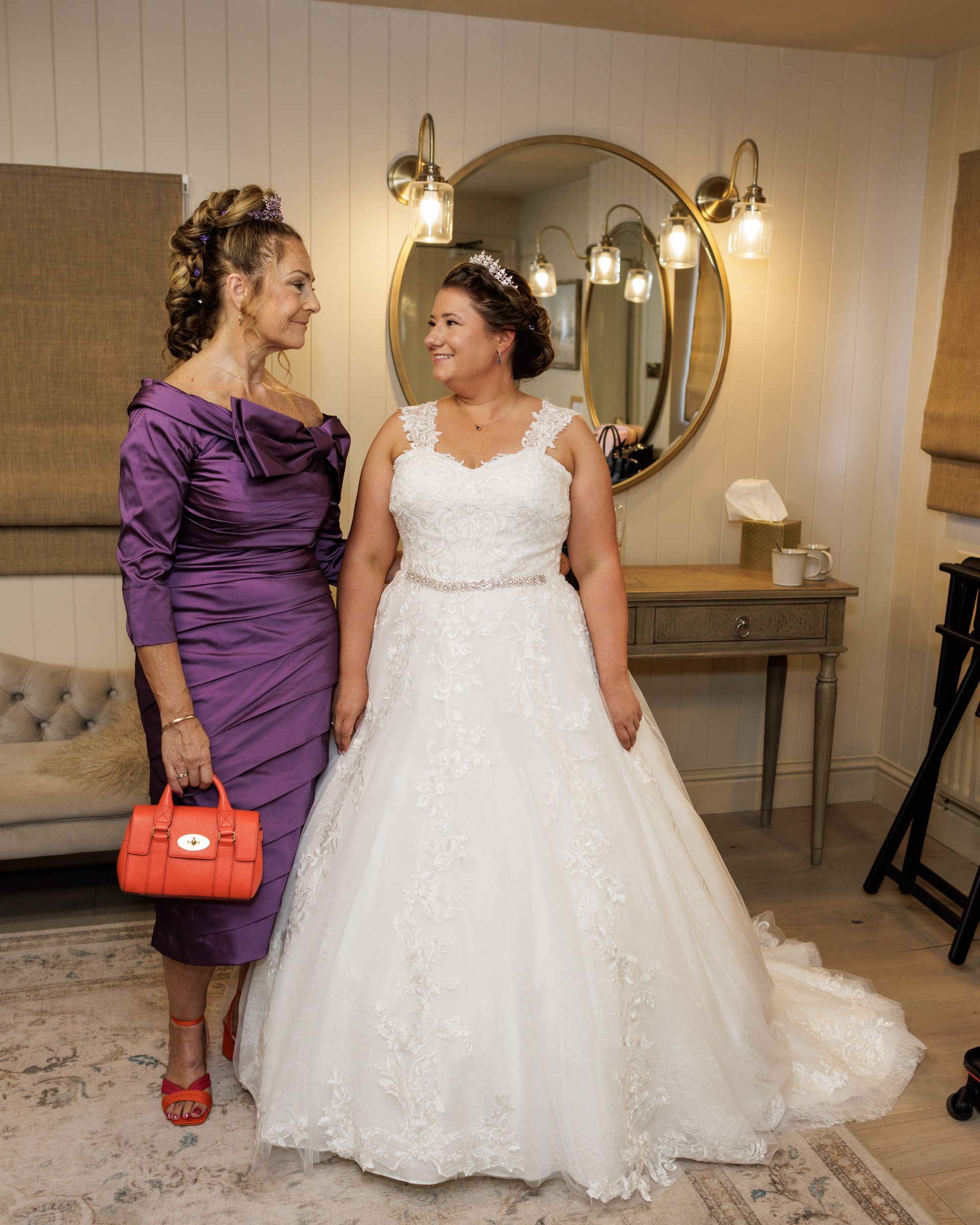 Bride in white lace ball gown stands with woman in purple satin dress holding red handbag in elegant bridal suite.