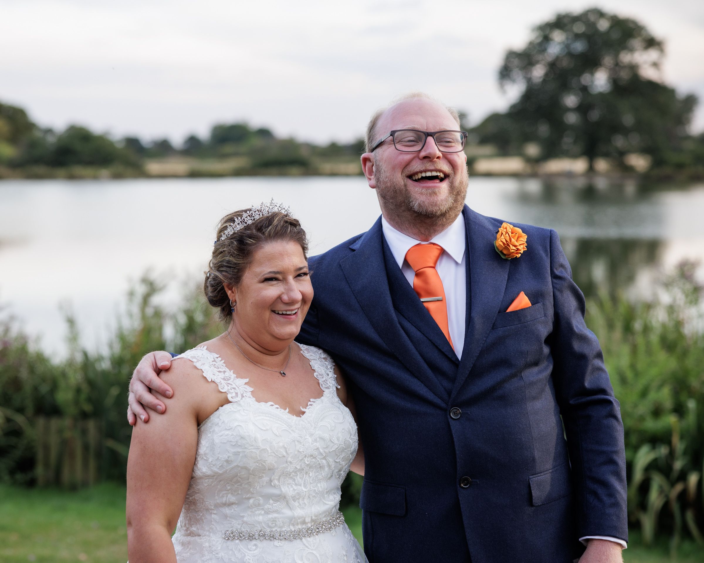 Happy bride and groom smiling outdoors by a lake, groom in navy suit with orange tie and boutonniere.