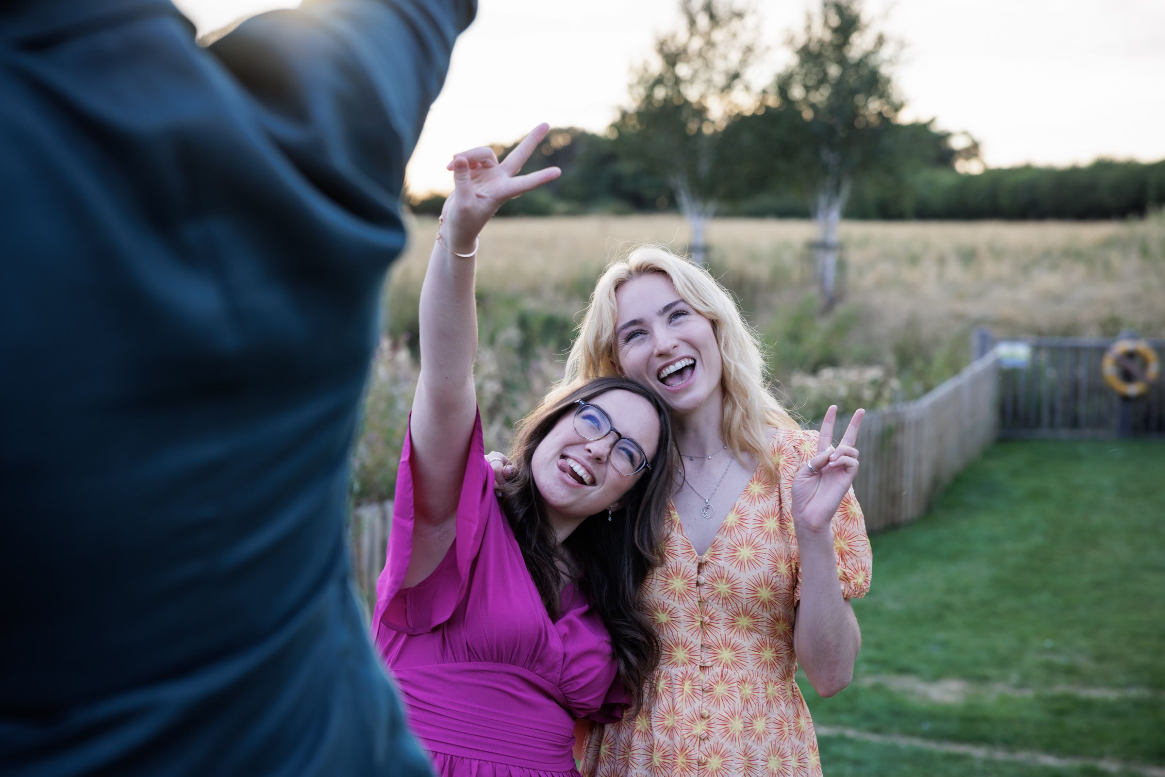 Two women smiling and posing outdoors, one in pink dress making peace sign, photographed at golden hour.