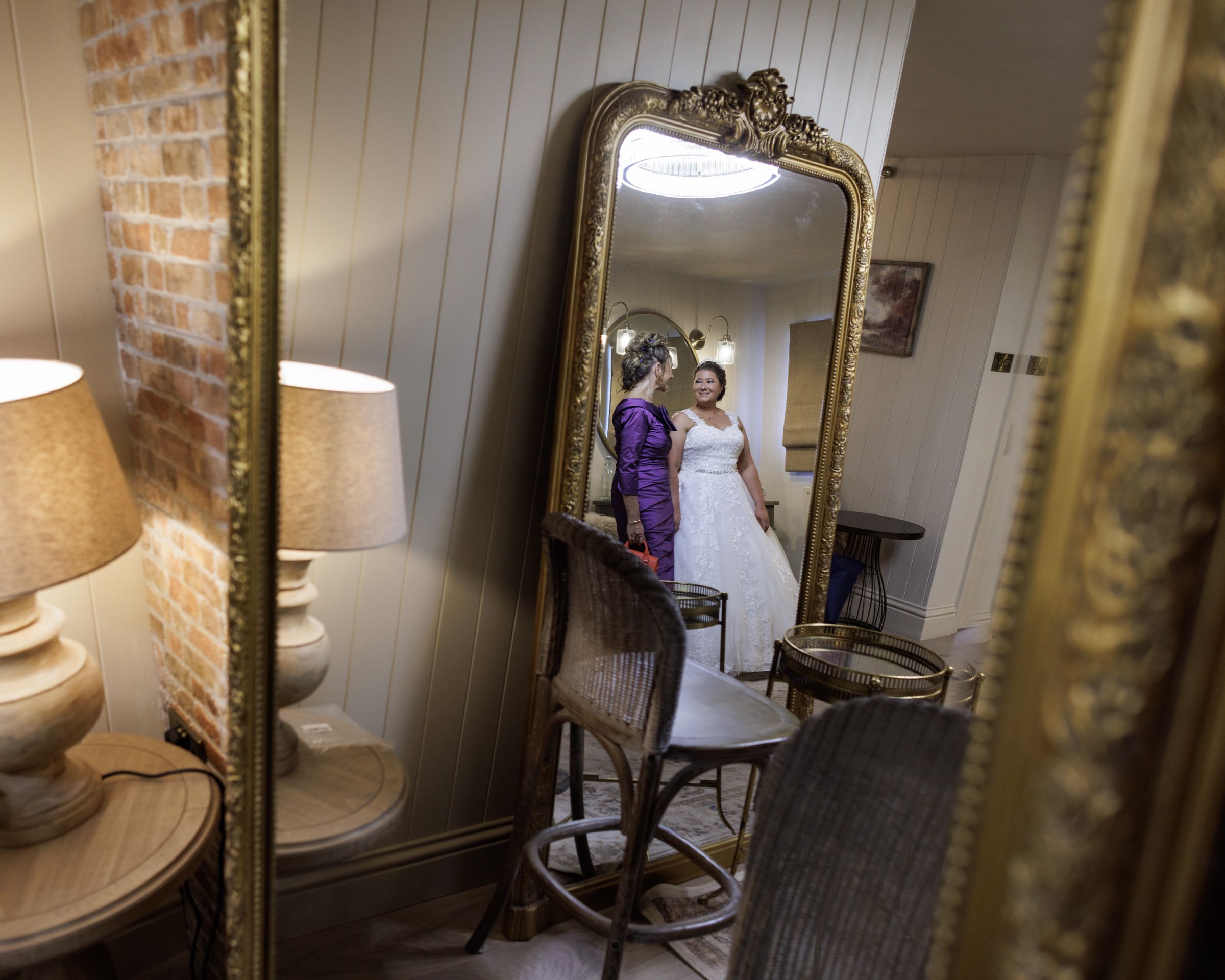 Bride in white gown reflected in ornate gold mirror during wedding preparations in elegant rustic venue.