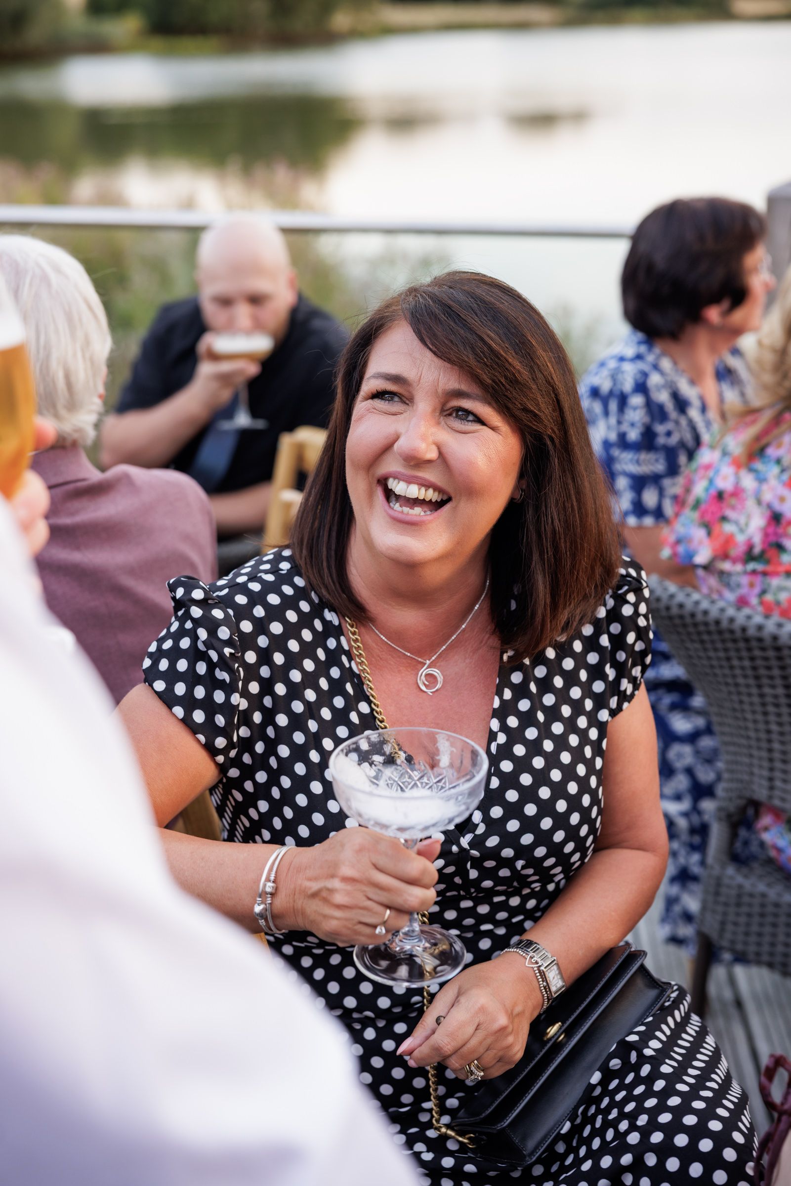 Woman in polka dot dress laughing at outdoor waterside gathering, holding cocktail glass.