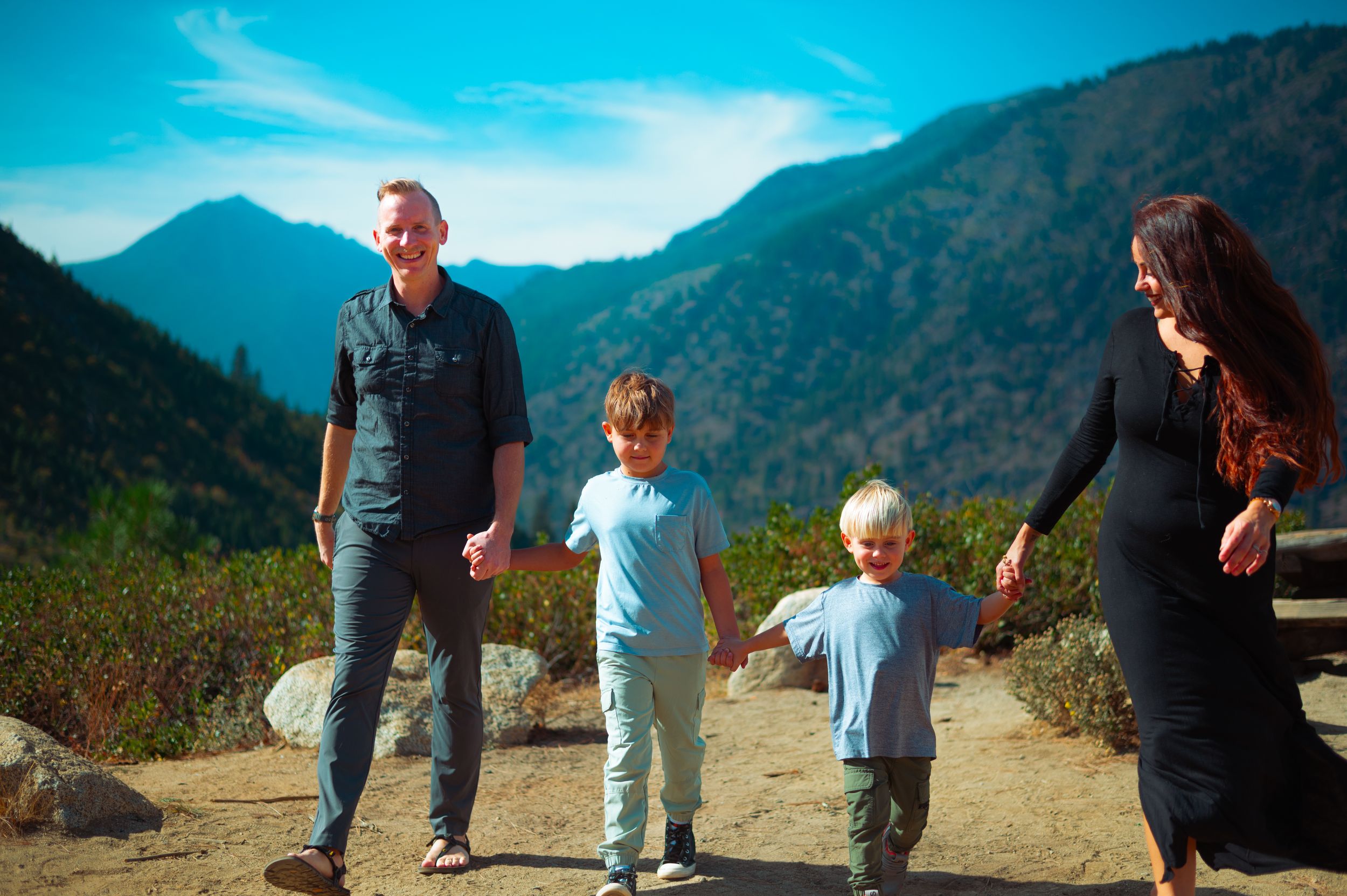 A happy family of four holds hands on a mountain trail surrounded by lush green hills and blue sky.