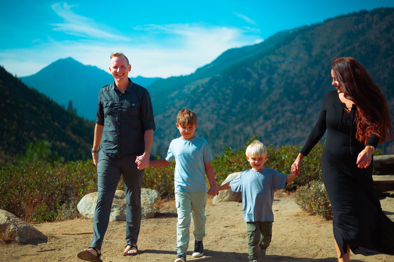 A happy family of four holds hands on a mountain trail surrounded by lush green hills and blue sky.