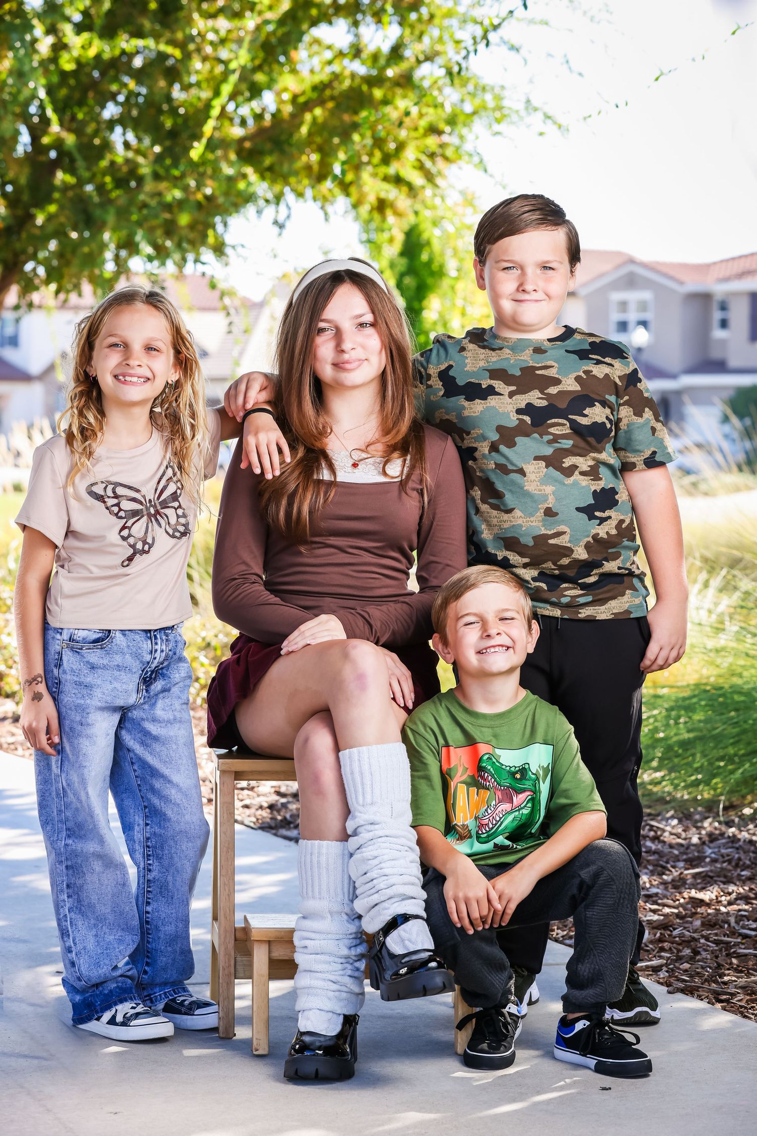 Four children posing outdoors near a suburban home, smiling together for a family portrait on a sunny day.