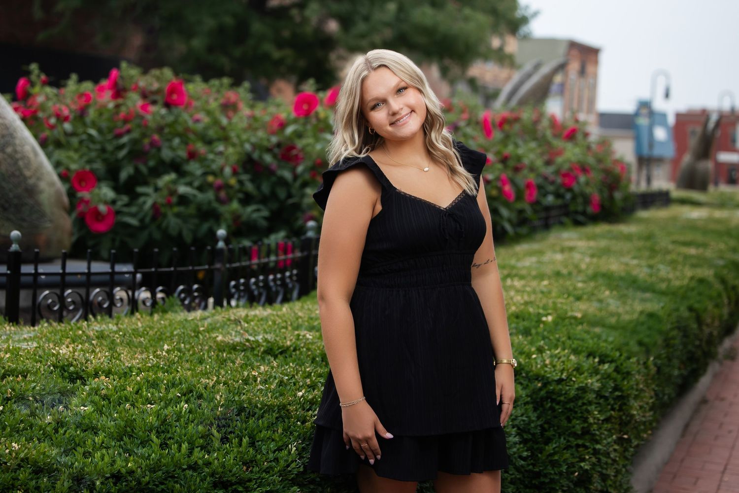 Young blonde woman in black dress smiling outdoors with red roses garden background.