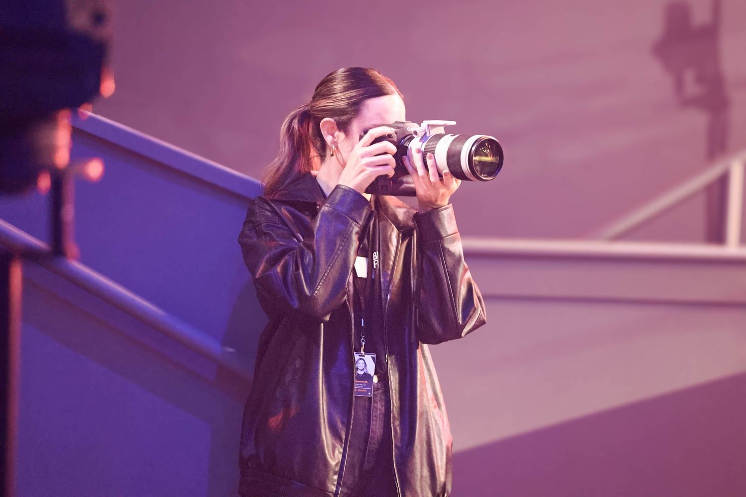 Woman in black jacket photographing with a professional camera at an indoor event with purple lighting.