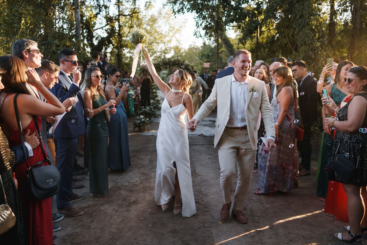 Newlyweds celebrate their outdoor wedding exit as guests cheer and line the path in warm evening light.