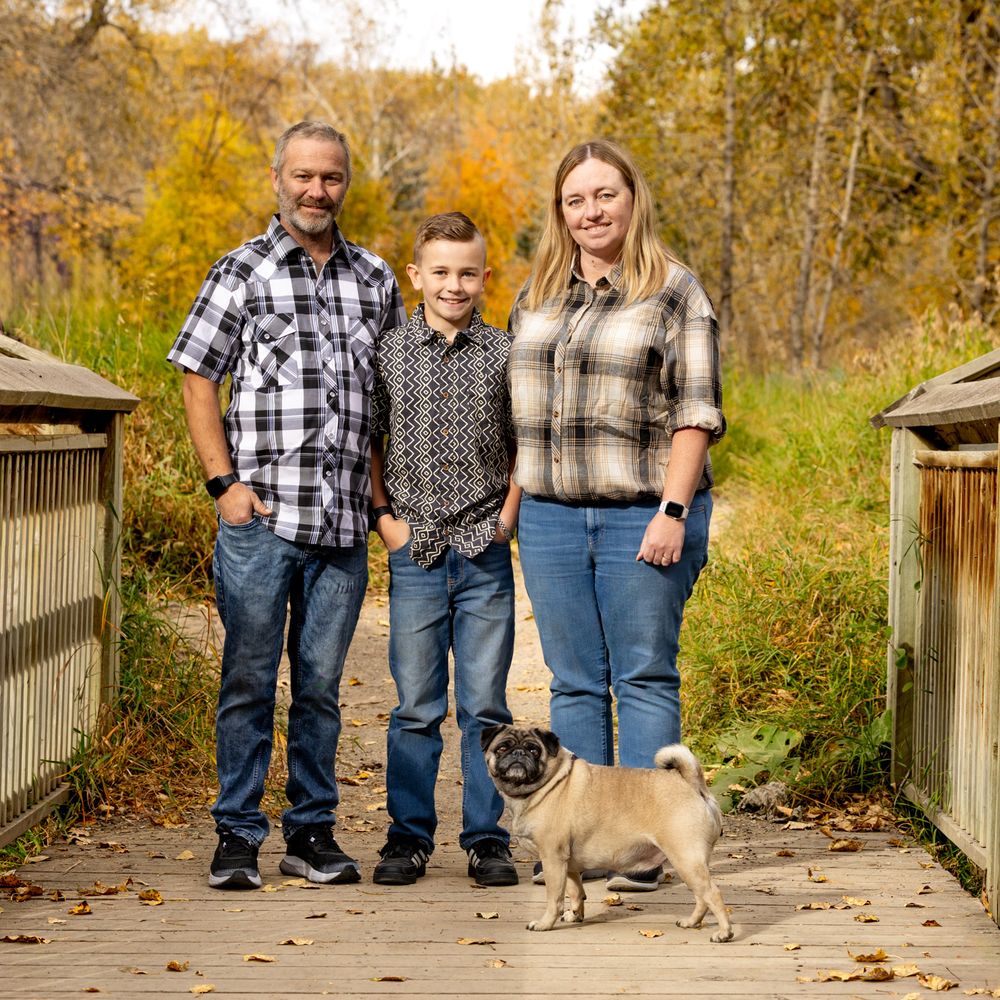 Family of three posing on a wooden bridge in autumn with a pug dog, surrounded by colorful fall foliage.