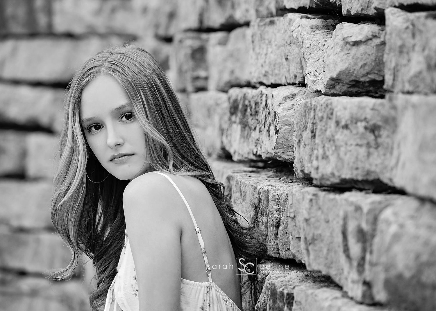 Black and white portrait of a young girl leaning against a stone wall, with long hair and a delicate dress.
