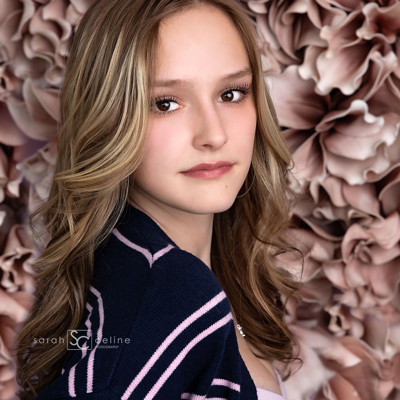 A young girl with wavy highlighted hair poses in a navy jacket against a soft pink floral backdrop.