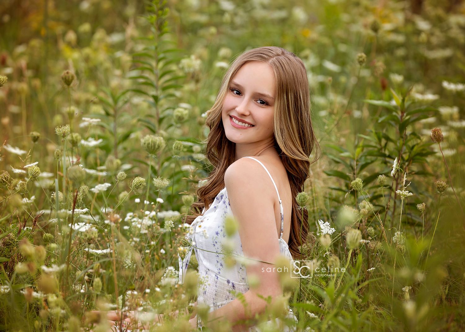 A smiling teenage girl in a white dress poses among wildflowers in a lush green field for senior portraits.