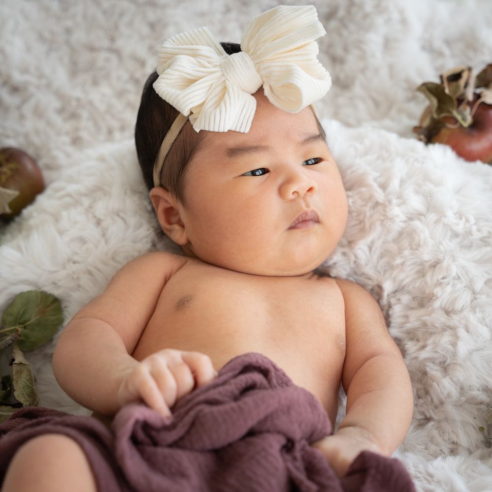 Awake newborn girl wearing a cream bow and lying on a soft blanket with autumn details.