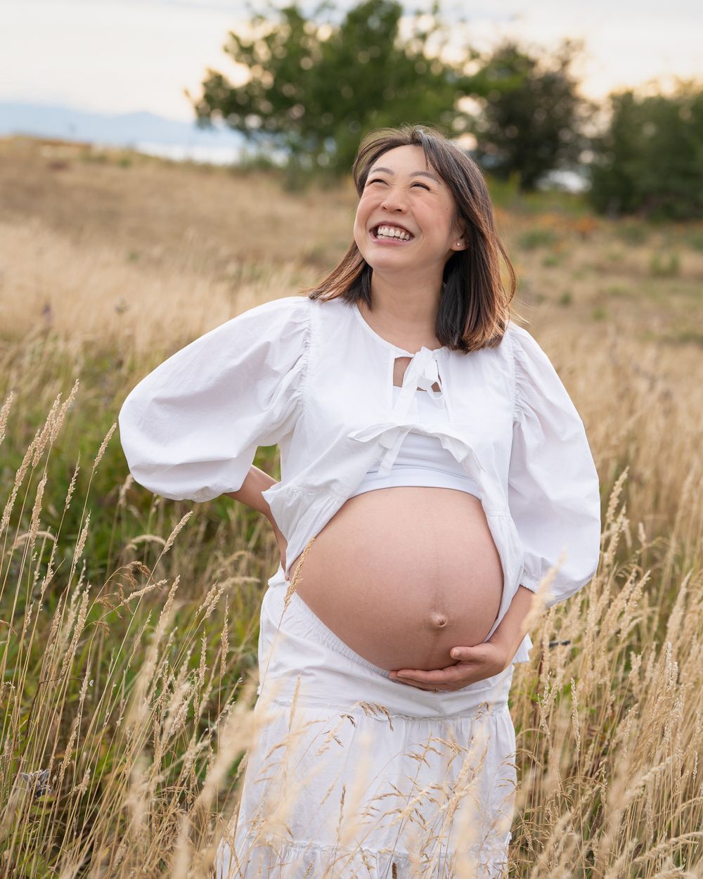 Pregnant woman smiling while standing in tall grass outdoors.