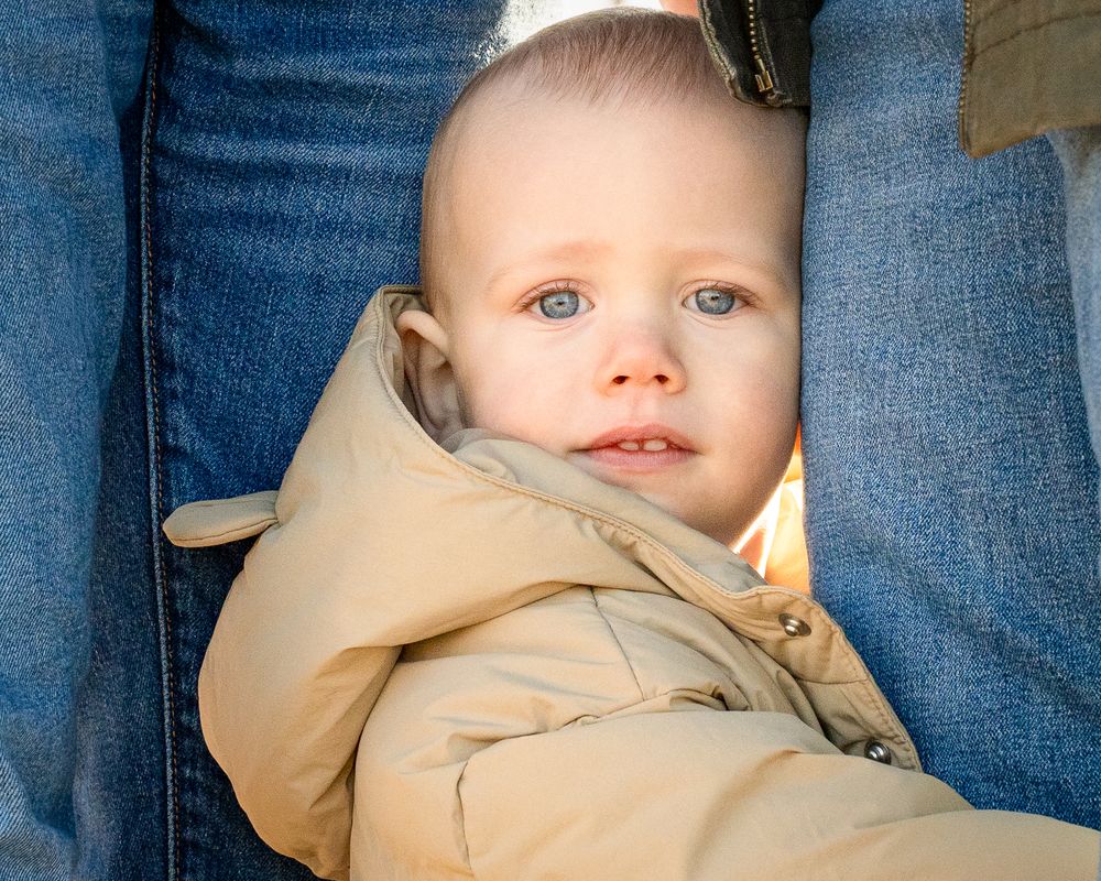 One-year-old child standing between a parent’s legs while wearing a tan winter coat.