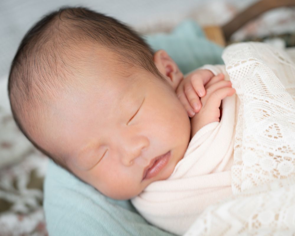 Sleeping newborn wrapped in cream fabric and resting under a lace blanket.