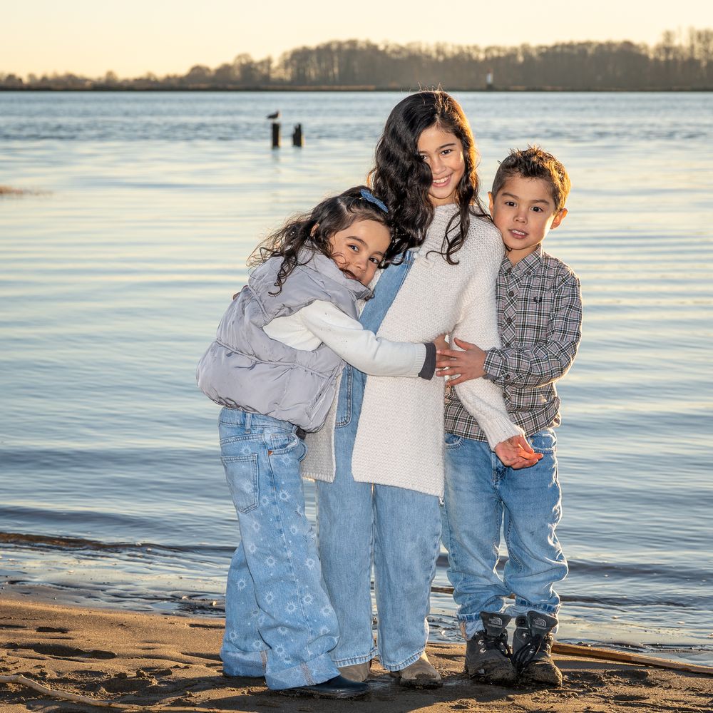 Three siblings standing together and hugging on the beach at sunset.