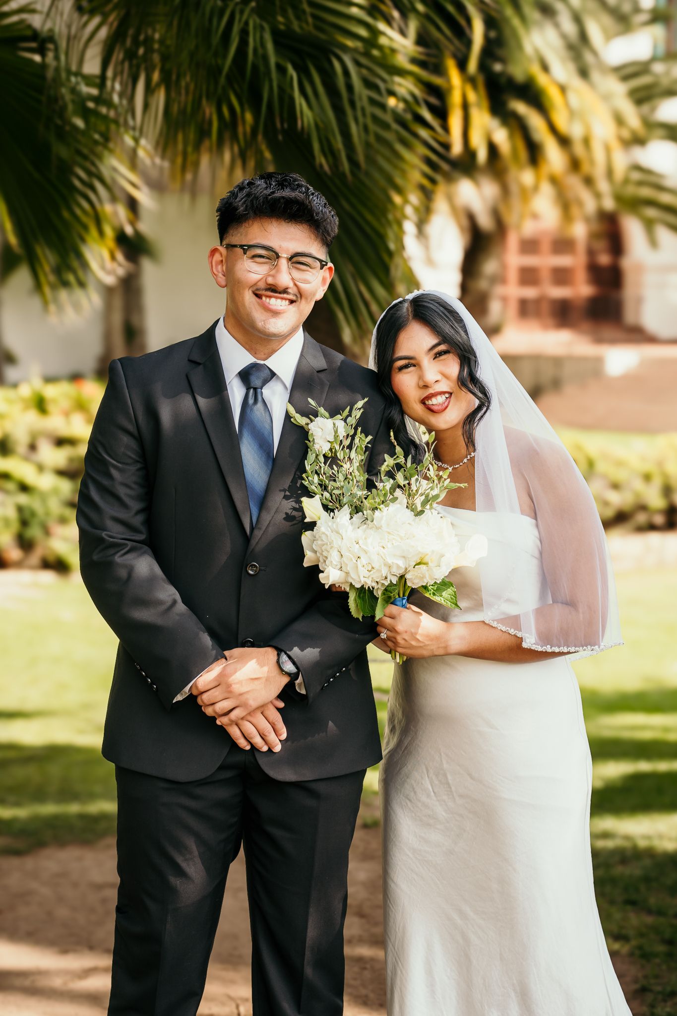 A smiling bride and groom pose outdoors in wedding attire, she holds a white bouquet, with palm trees in the background.