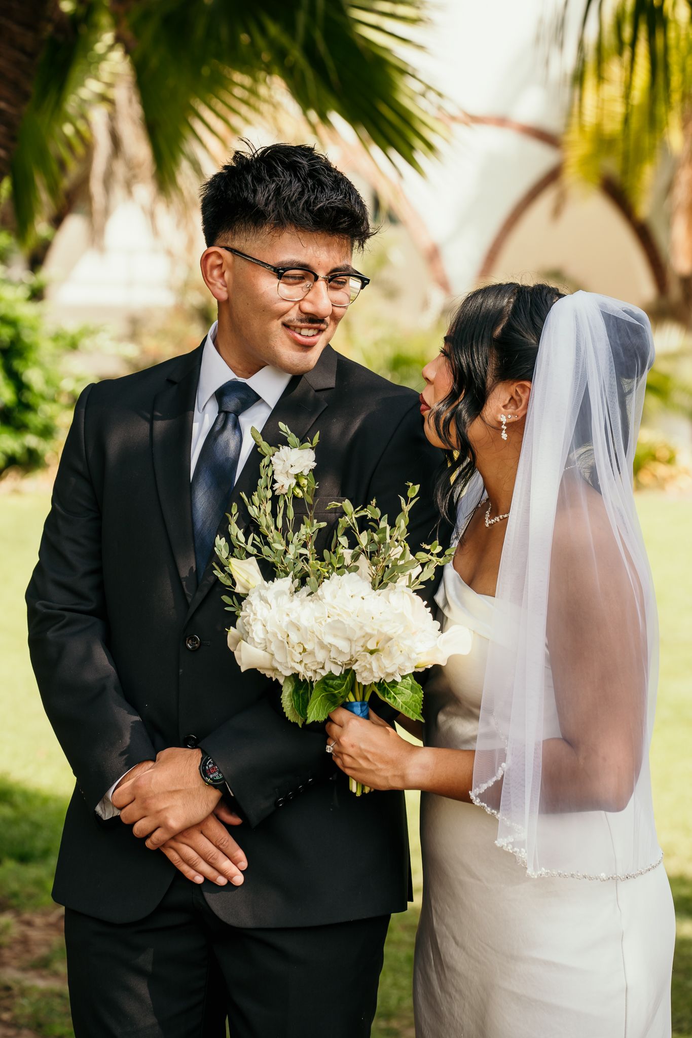 Smiling groom in black suit and glasses holds white floral bouquet beside bride in white dress and veil outdoors.
