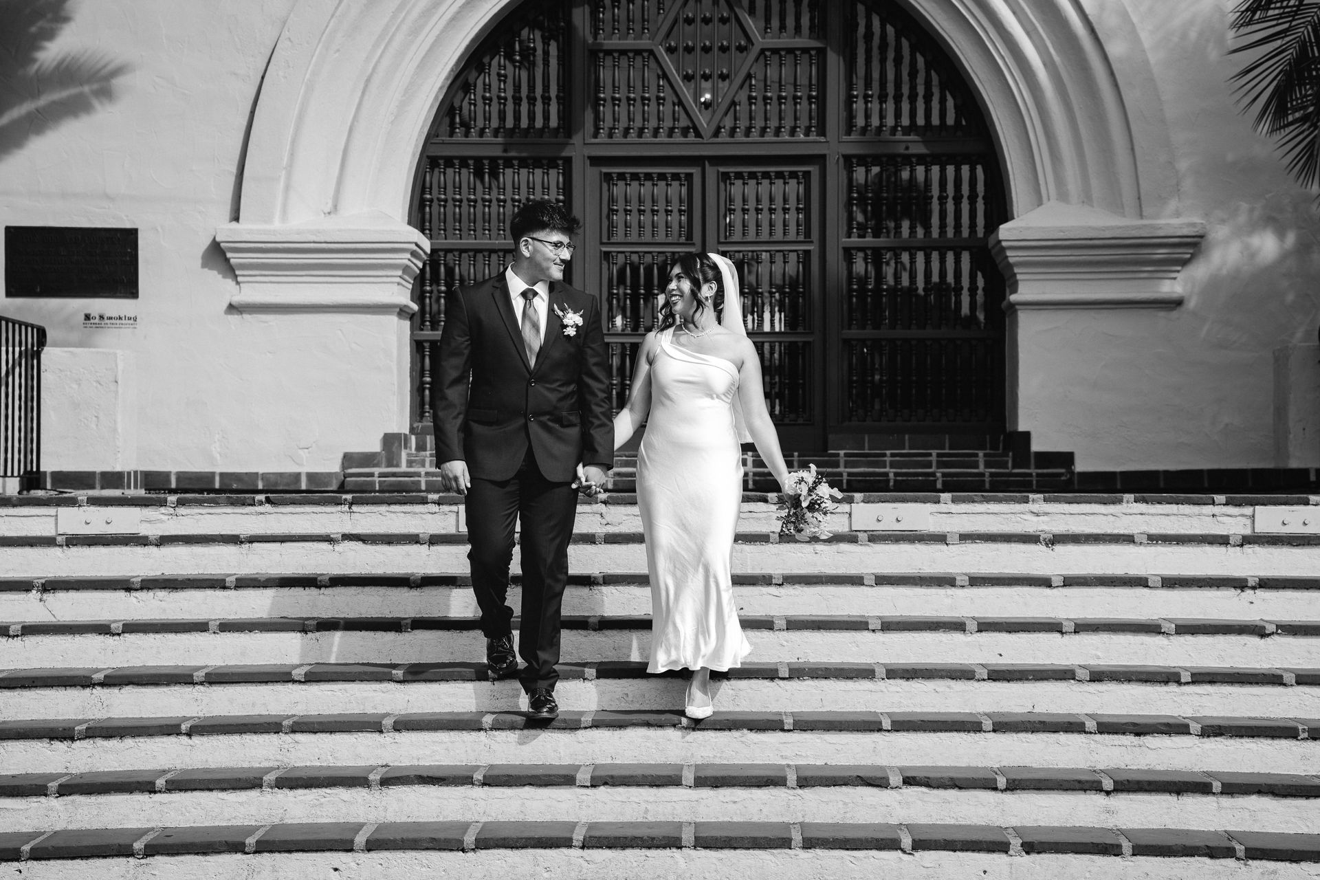 Black and white photo of a bride and groom holding hands on church steps after their wedding ceremony.