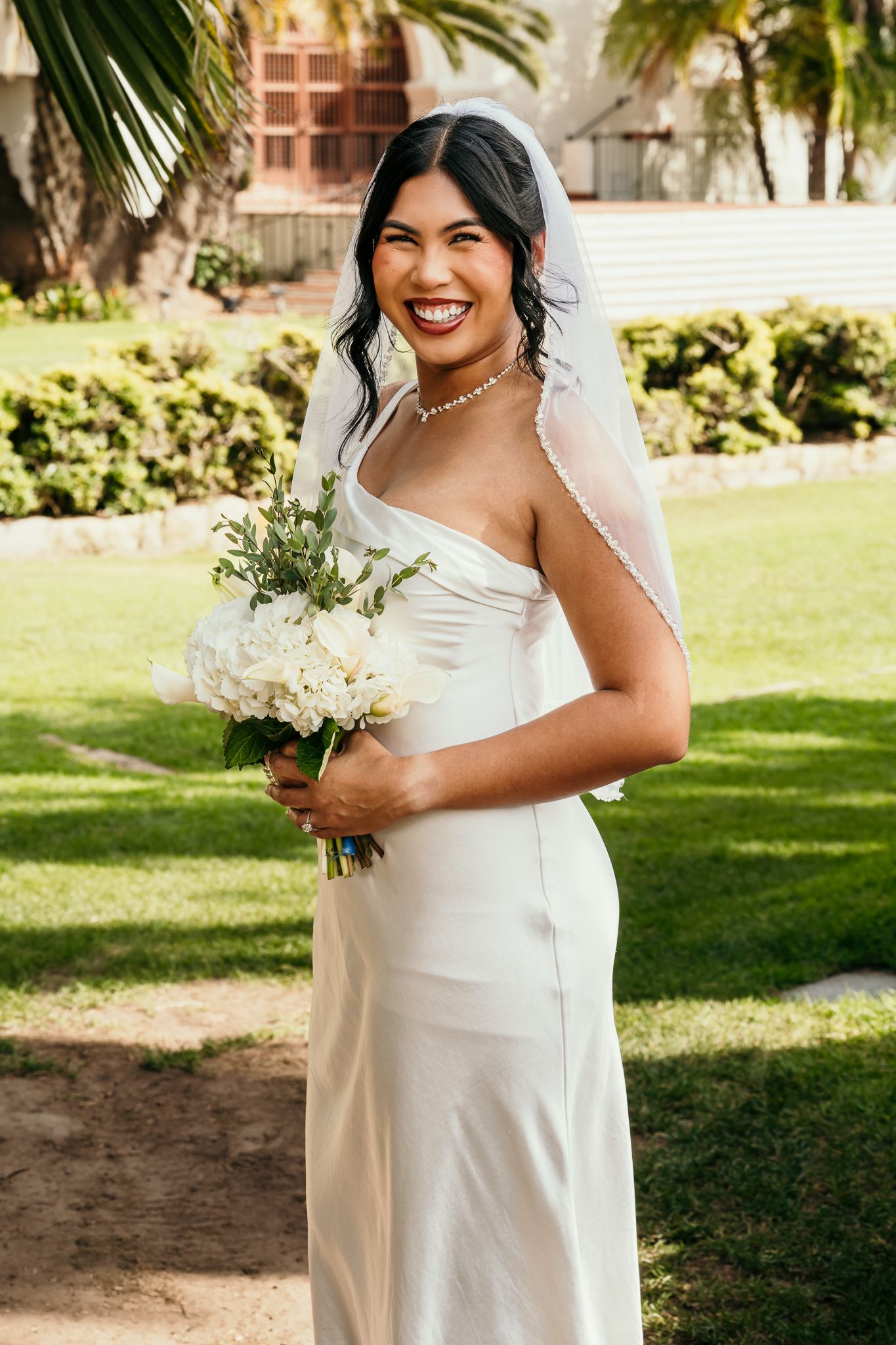 Smiling bride in white satin gown and veil holds white bouquet outdoors on green lawn.