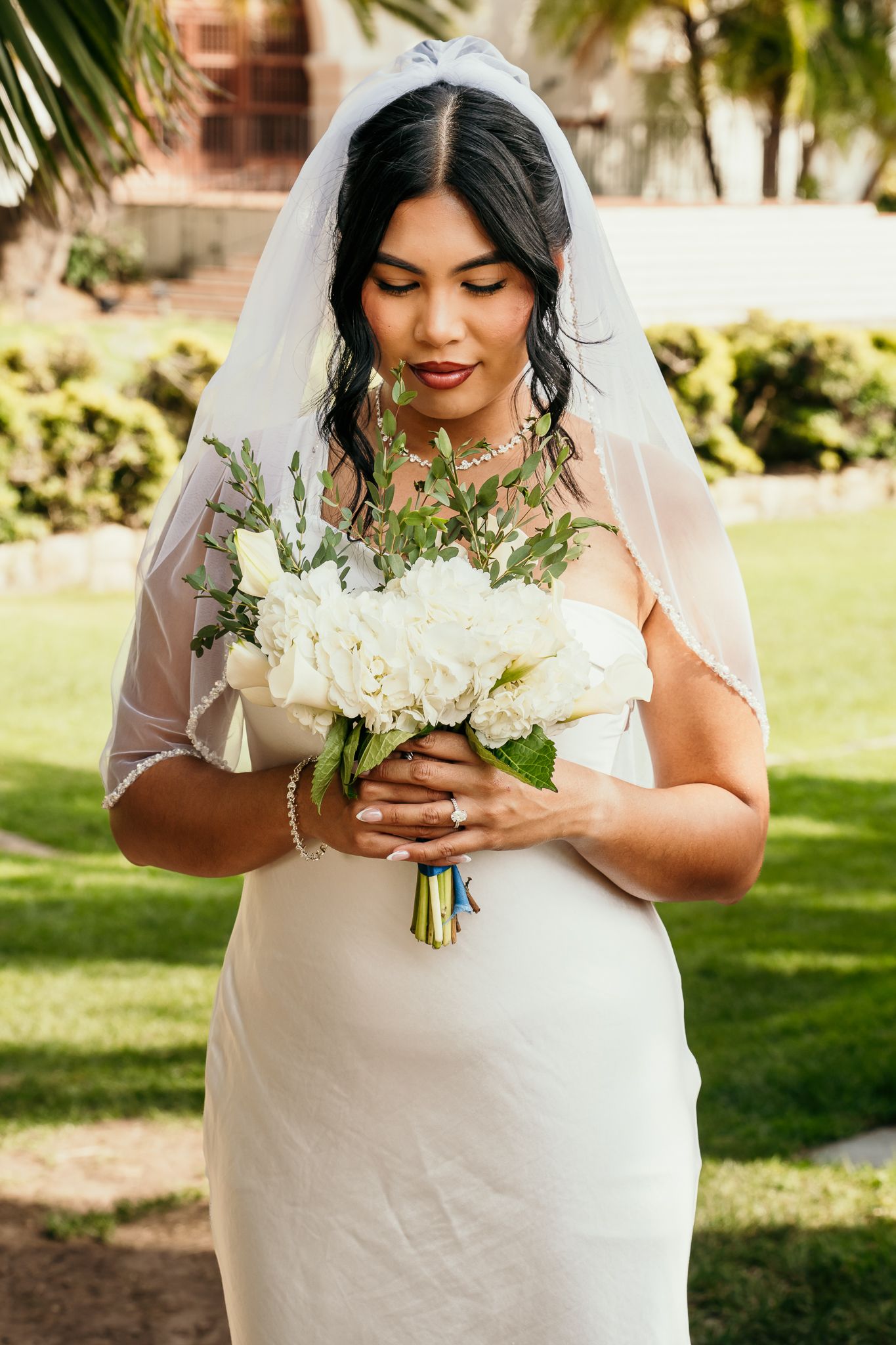 Bride in white gown and veil holds white floral bouquet outdoors on green lawn.