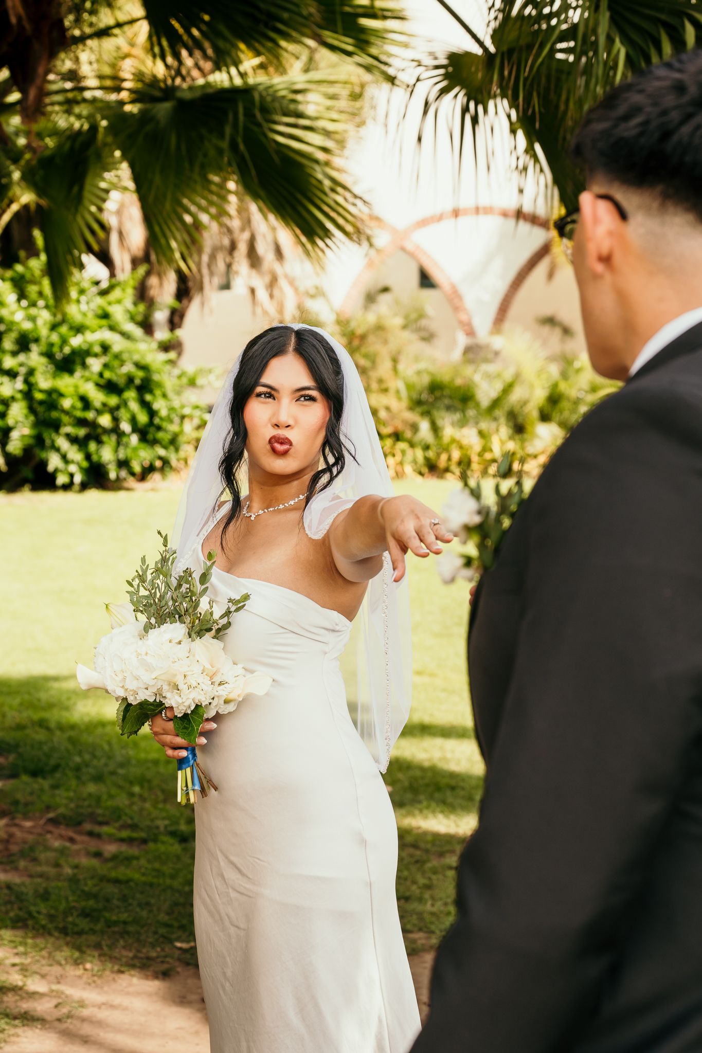 Bride in white gown holding bouquet points at groom outdoors near tropical palm trees.