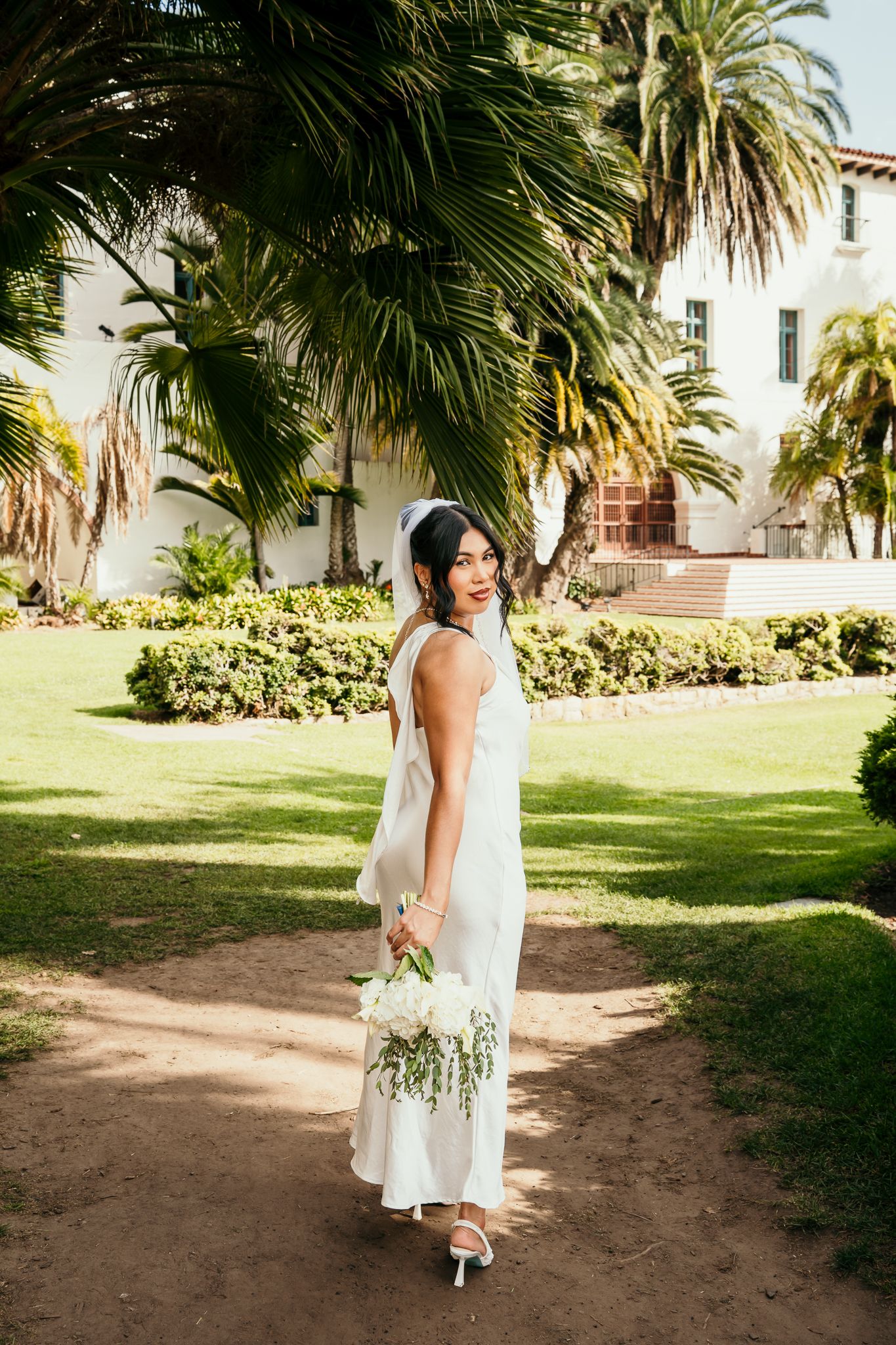 Bride in white dress holding bouquet poses in lush garden with palm trees and white building backdrop.