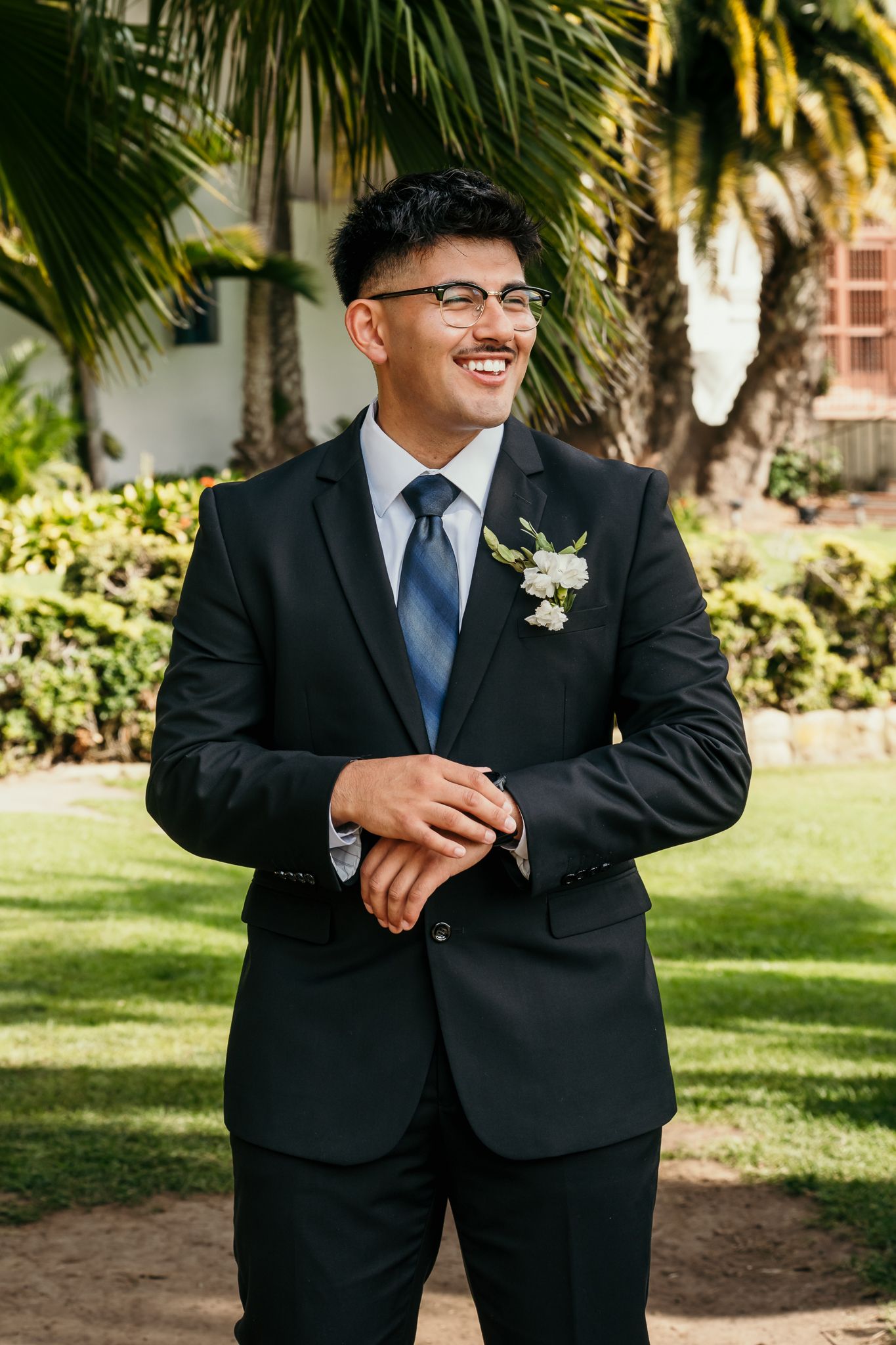 Smiling groom in black suit with boutonniere adjusts cufflinks outdoors near palm trees.
