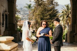 Outdoor wedding ceremony with bride in white gown, officiant in navy dress reading vows, and groom in black suit.