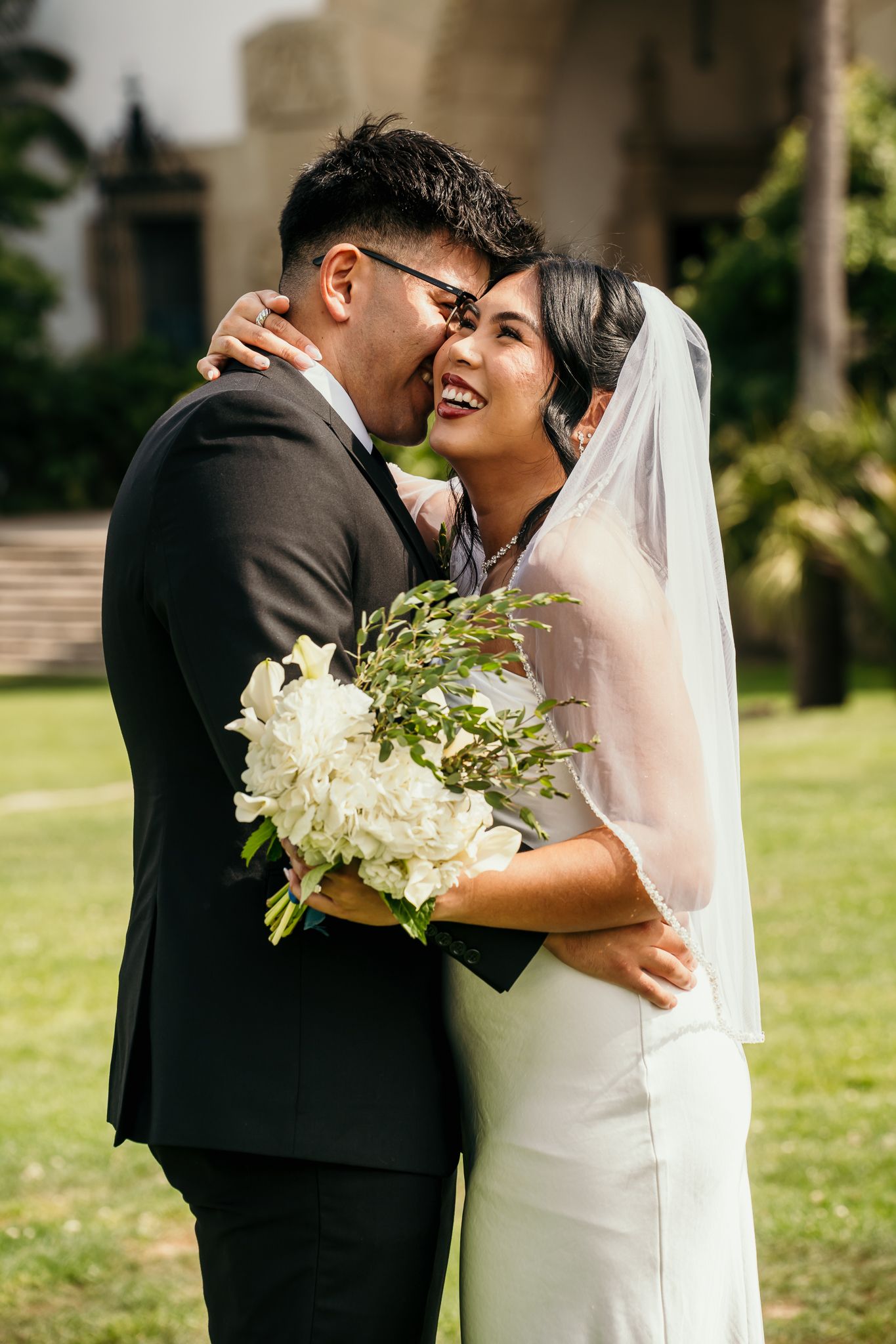 Smiling bride in white gown and veil holds floral bouquet as groom in black suit kisses her cheek outdoors.